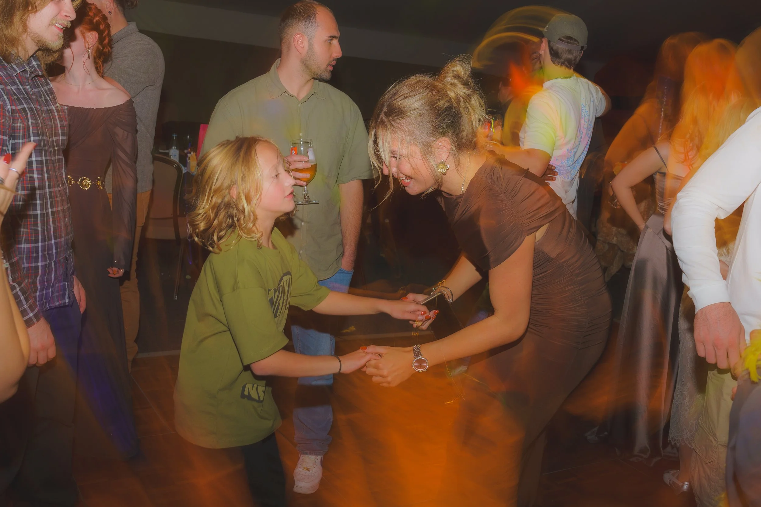 A group of people at a party, with two women in the center engaged in a conversation while one woman holds the hand of a girl, all smiling. Others are mingling in the background, with some holding drinks.