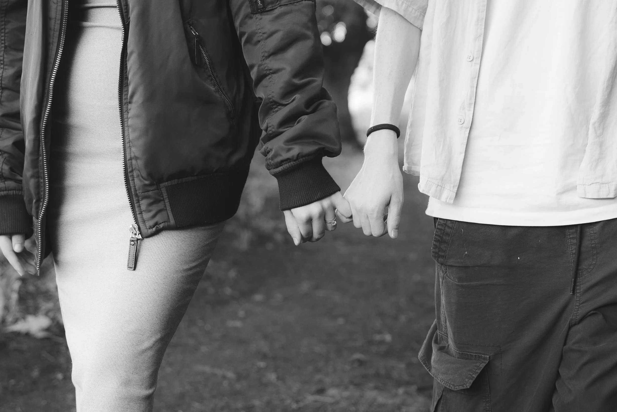 A close-up black-and-white photo of two people holding hands, showing only their torsos and hands. One person is wearing a dark bomber jacket and a light-colored top, and the other is wearing a light-colored shirt and dark jeans. They are outdoors, near some trees or bushes.
