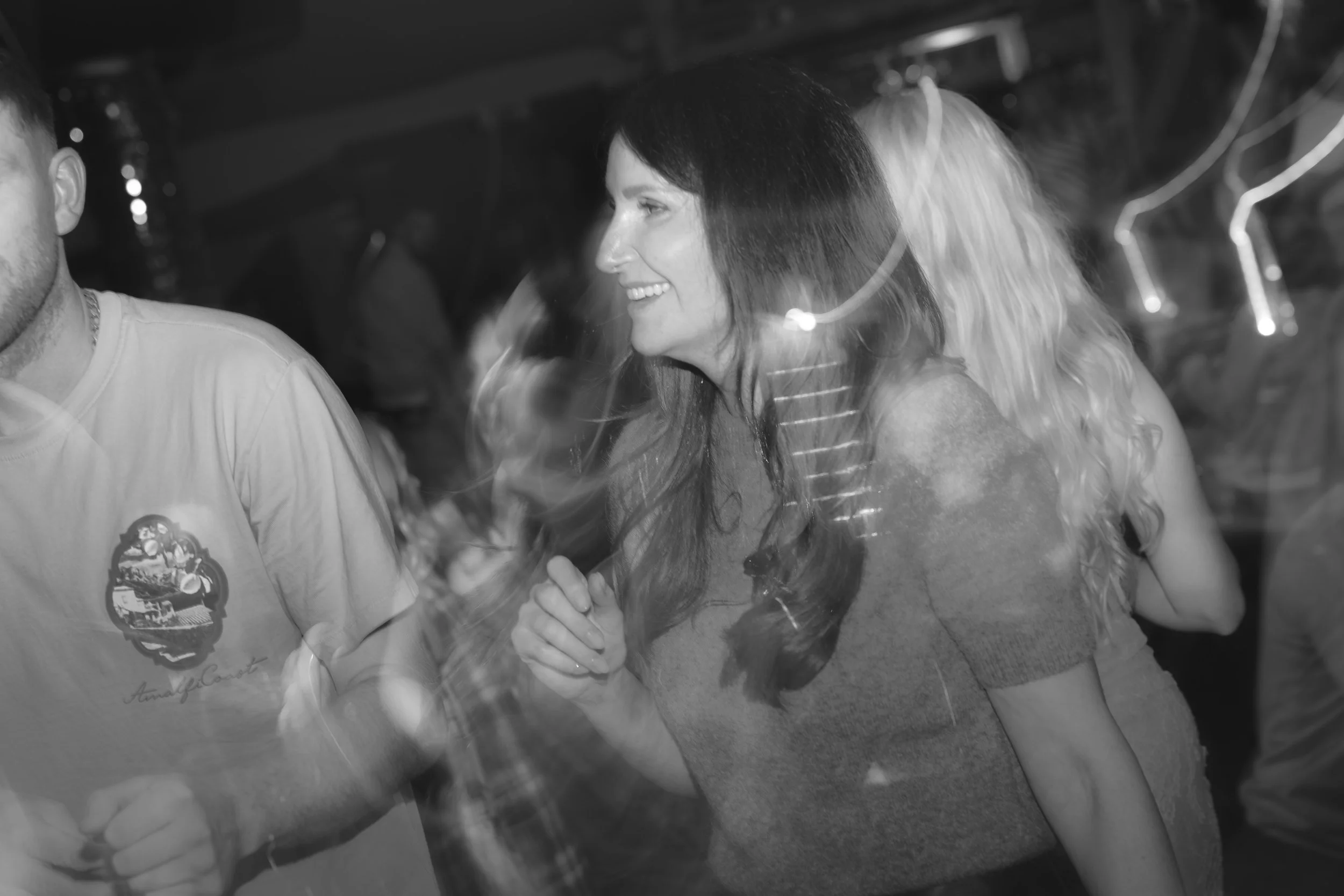 People dancing at a social gathering, including a smiling woman with long hair wearing a striped shirt. The scene is in black and white with some light streaks.