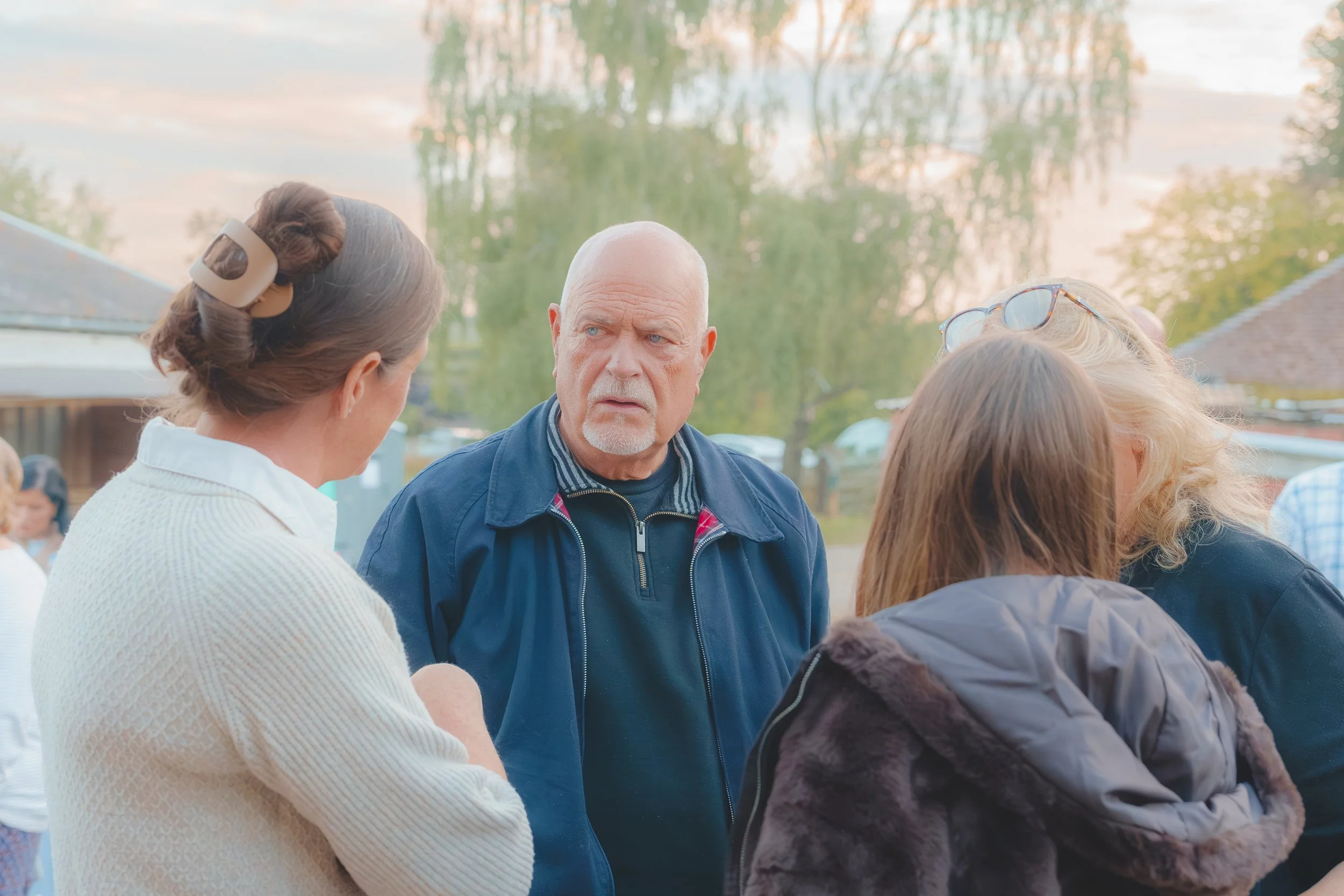 A group of people engaged in a conversation outdoors during sunset, with trees and rooftops in the background.