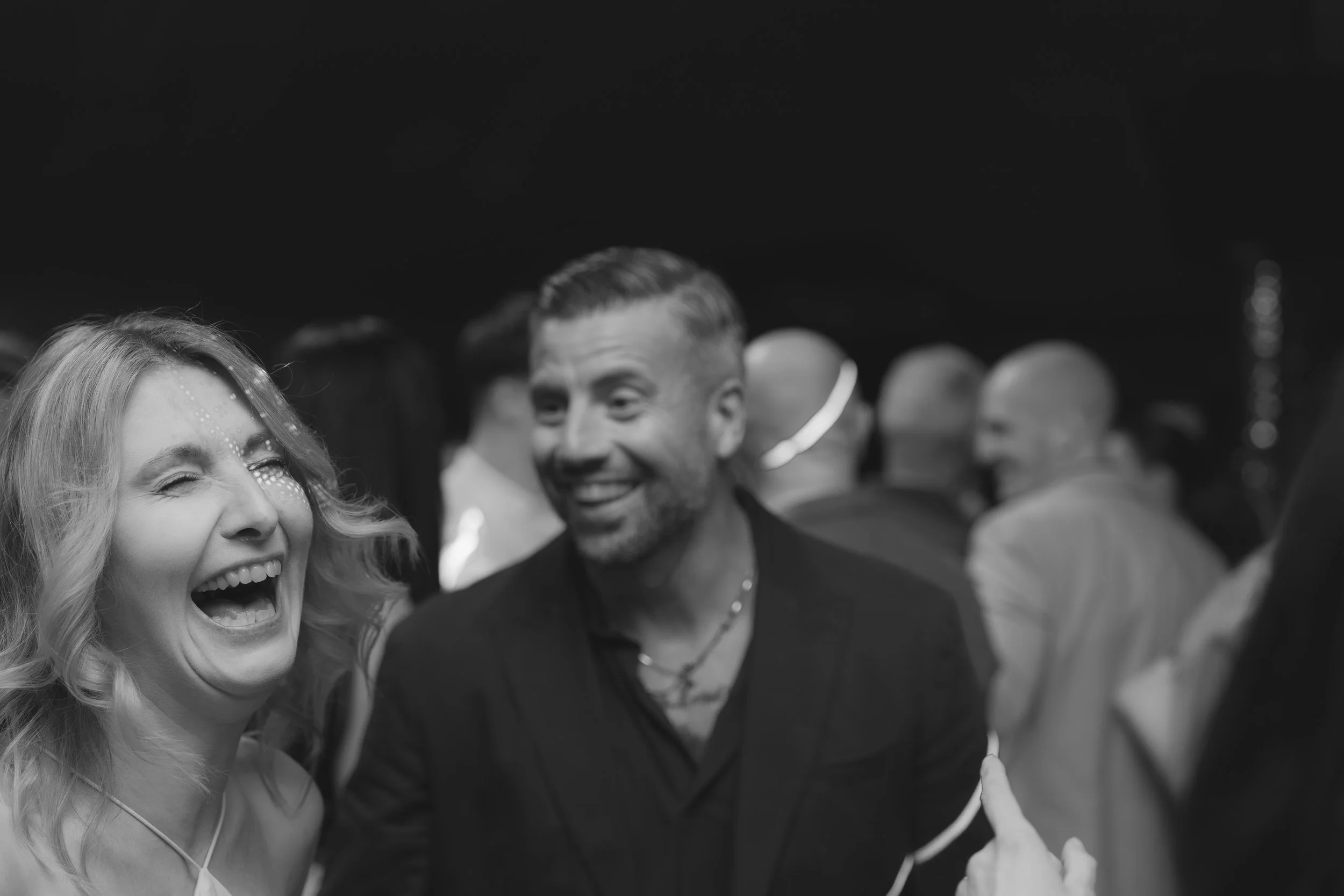 A black-and-white photo of a group of people at a celebration or party, with a woman laughing and a man smiling in the foreground.