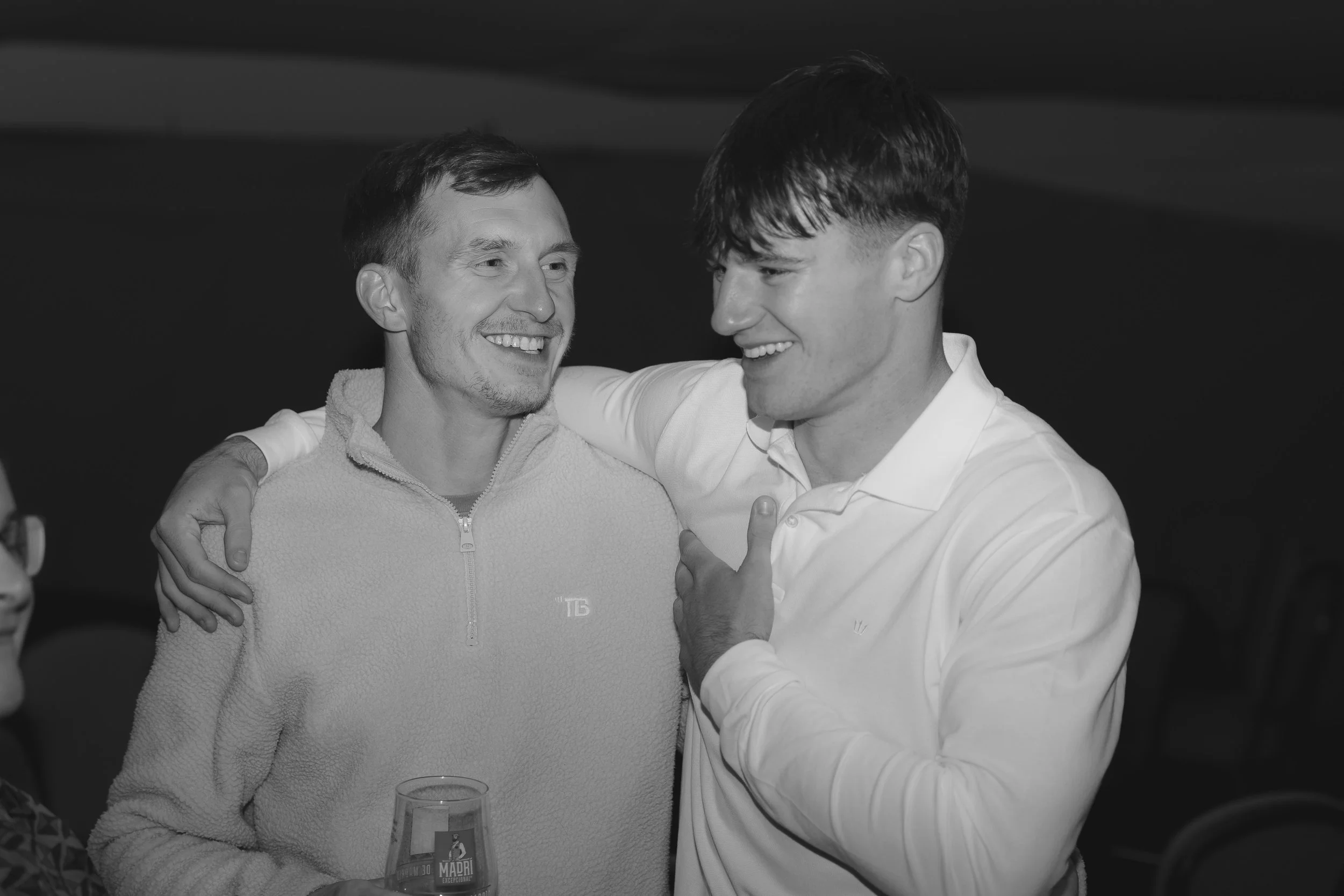 Two men are smiling and embracing at a social gathering. One man, with short dark hair, is wearing a light-colored zip-up sweater, and the other, with slightly longer dark hair, is dressed in a white shirt. There is a glass of beer on the table in fr