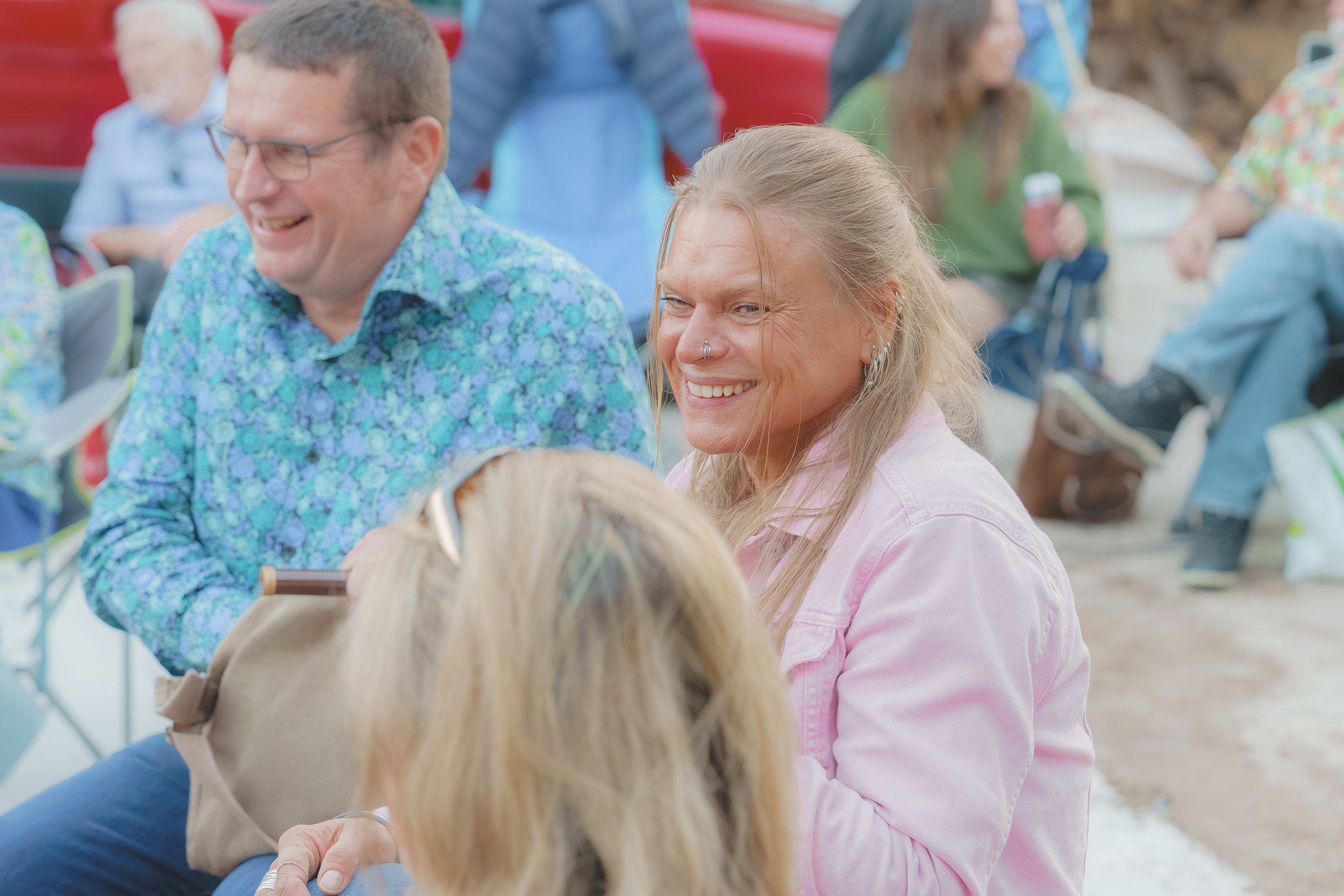 Several people, including a woman with a nose ring and a man with glasses, are outdoors at a gathering, smiling and talking.