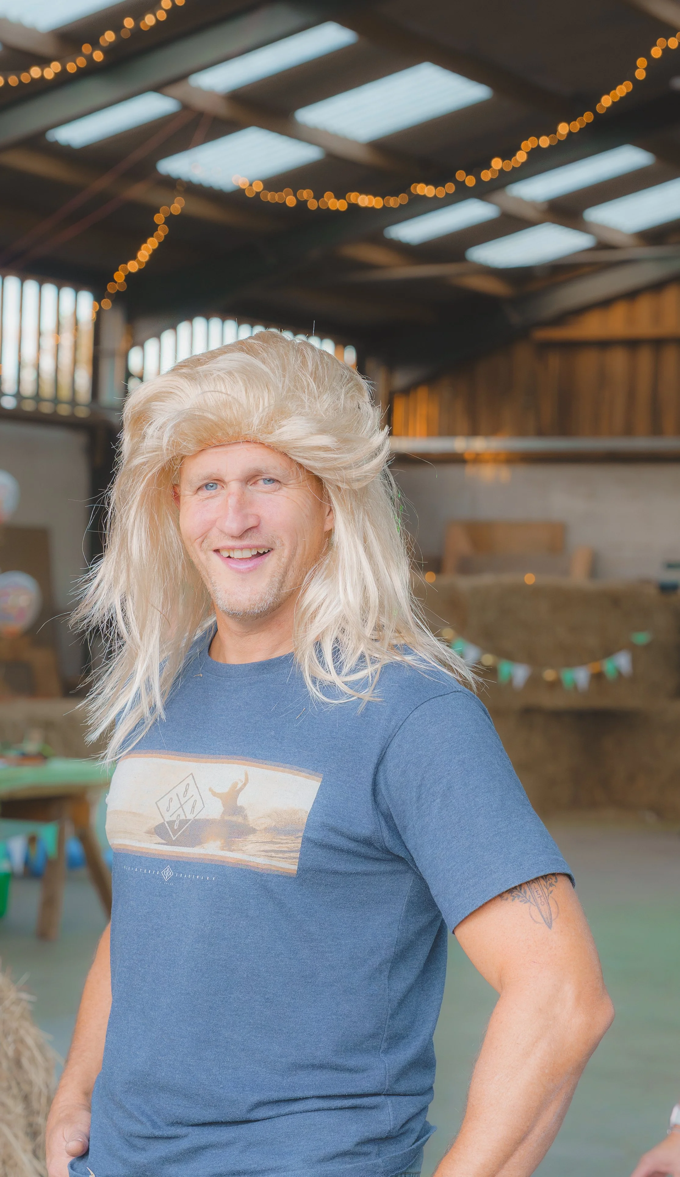 A smiling man with long, platinum blonde hair and a tattoo on his left arm in an indoor barn decorated with string lights and bunting, wearing a blue T-shirt with a beach scene graphic.