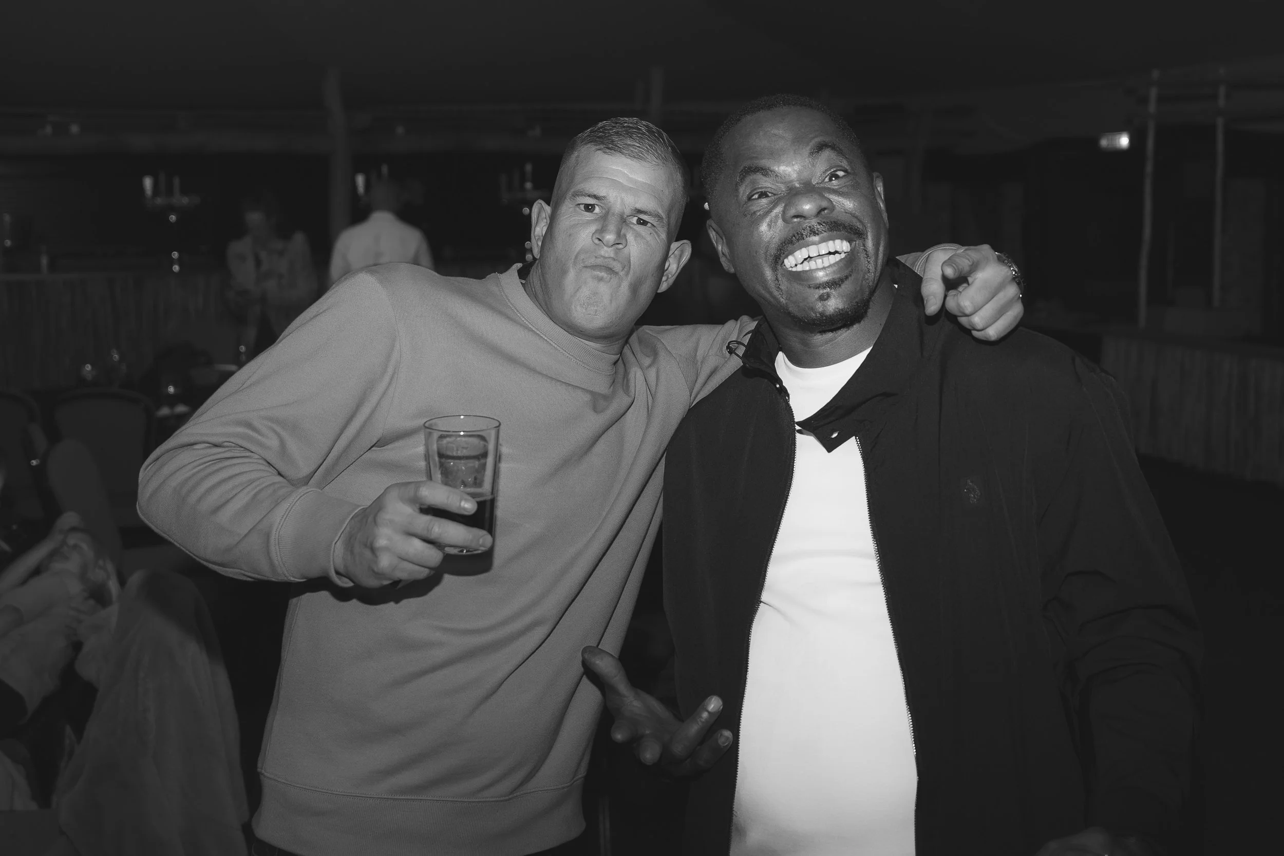 Two men smiling and posing together at a social gathering, one holding a drink, with a dark indoor background.