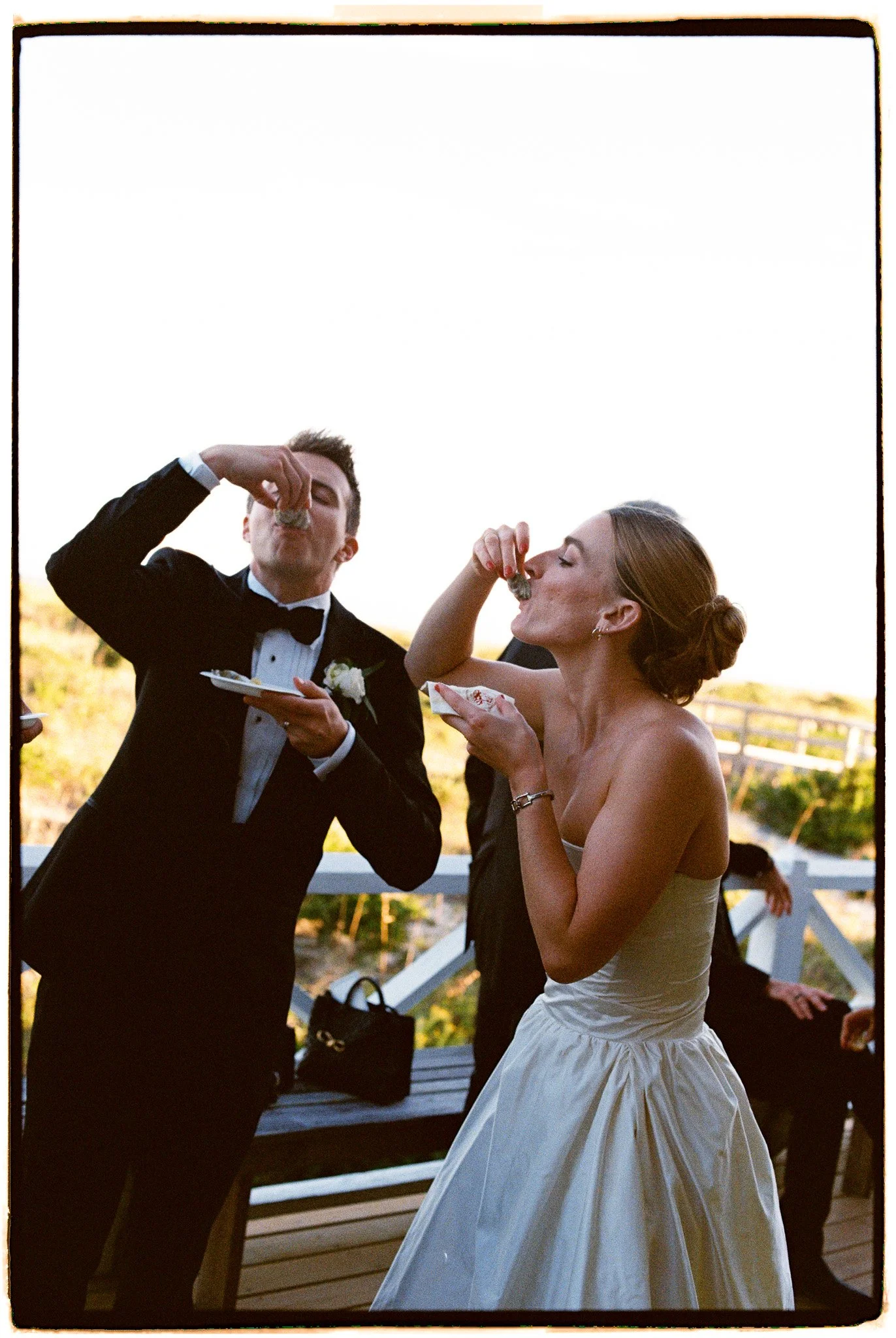 A man and woman in formal attire, eating cake at a celebration or wedding, outdoors during sunset.