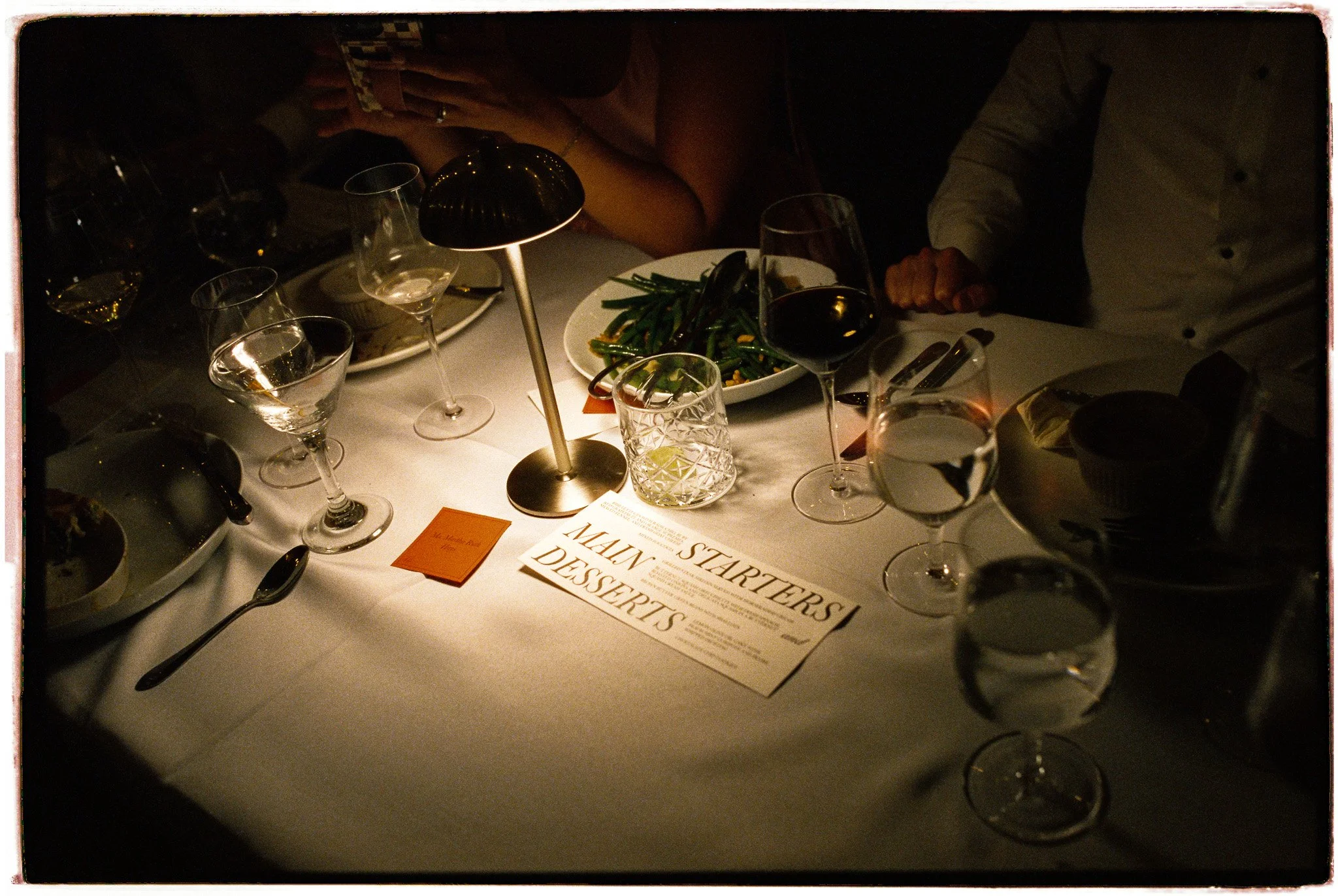 A dimly lit table set for dining with wine glasses, plates, silverware, a bowl of green beans, and a small lamp in the center, with people sitting around it.