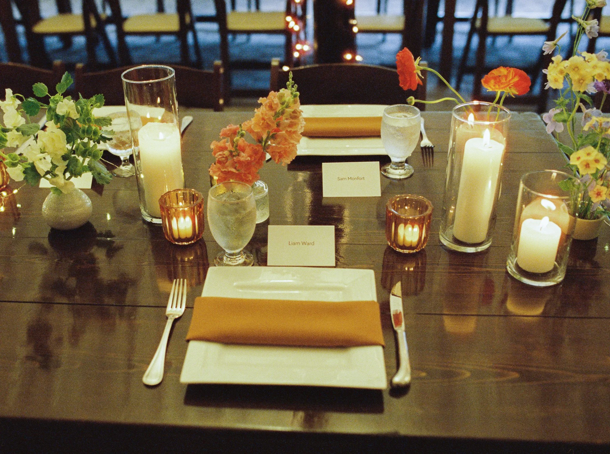 A dining table set for a formal event with candlelit decorations, floral arrangements, place cards, and cutlery, with chairs in the background.
