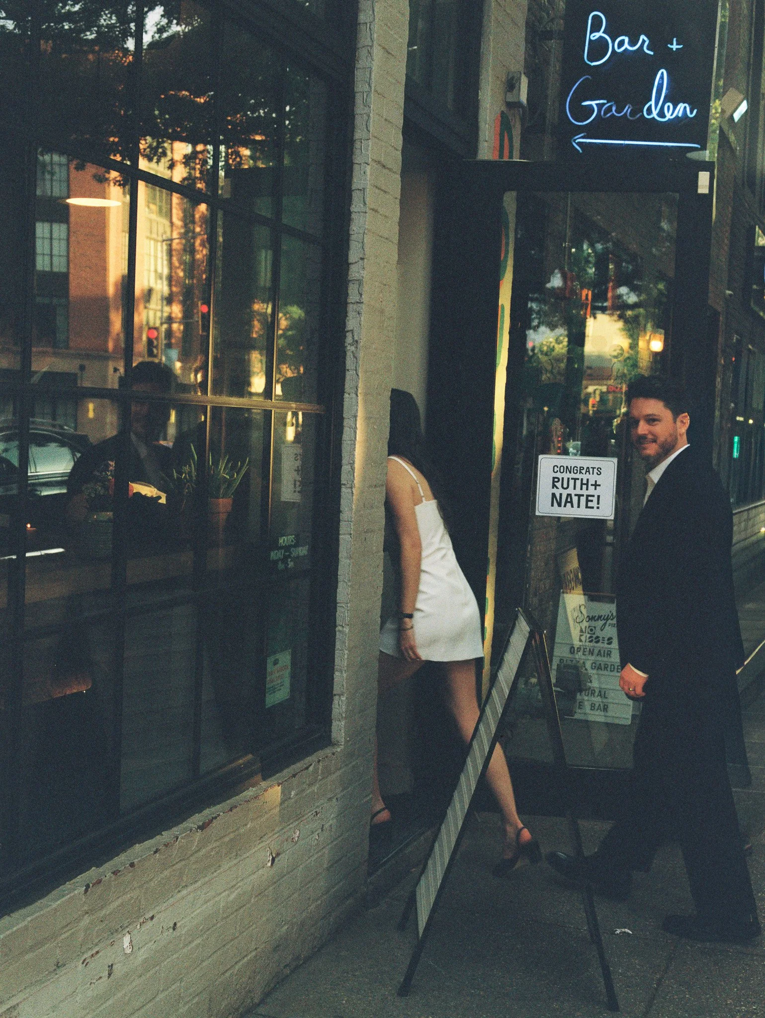 A man in a black suit and a woman in a white dress are standing outside a storefront. There is a neon sign that reads 'Bar + Garden' with an arrow pointing to the left. A board in front of the window says 'Congrats Ruth + Nate!' with additional text about food and the garden. The scene appears to be during sunset or early evening.