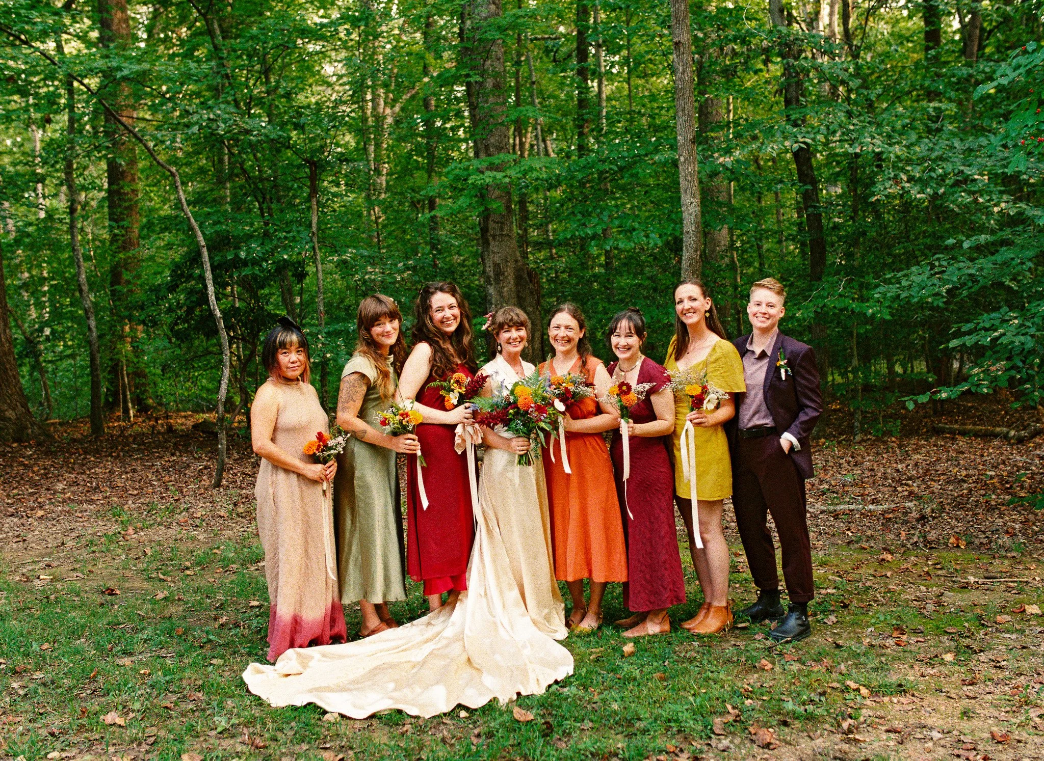 Group of women in colorful dresses holding bouquets, standing outdoors in a wooded area, smiling for a photo.