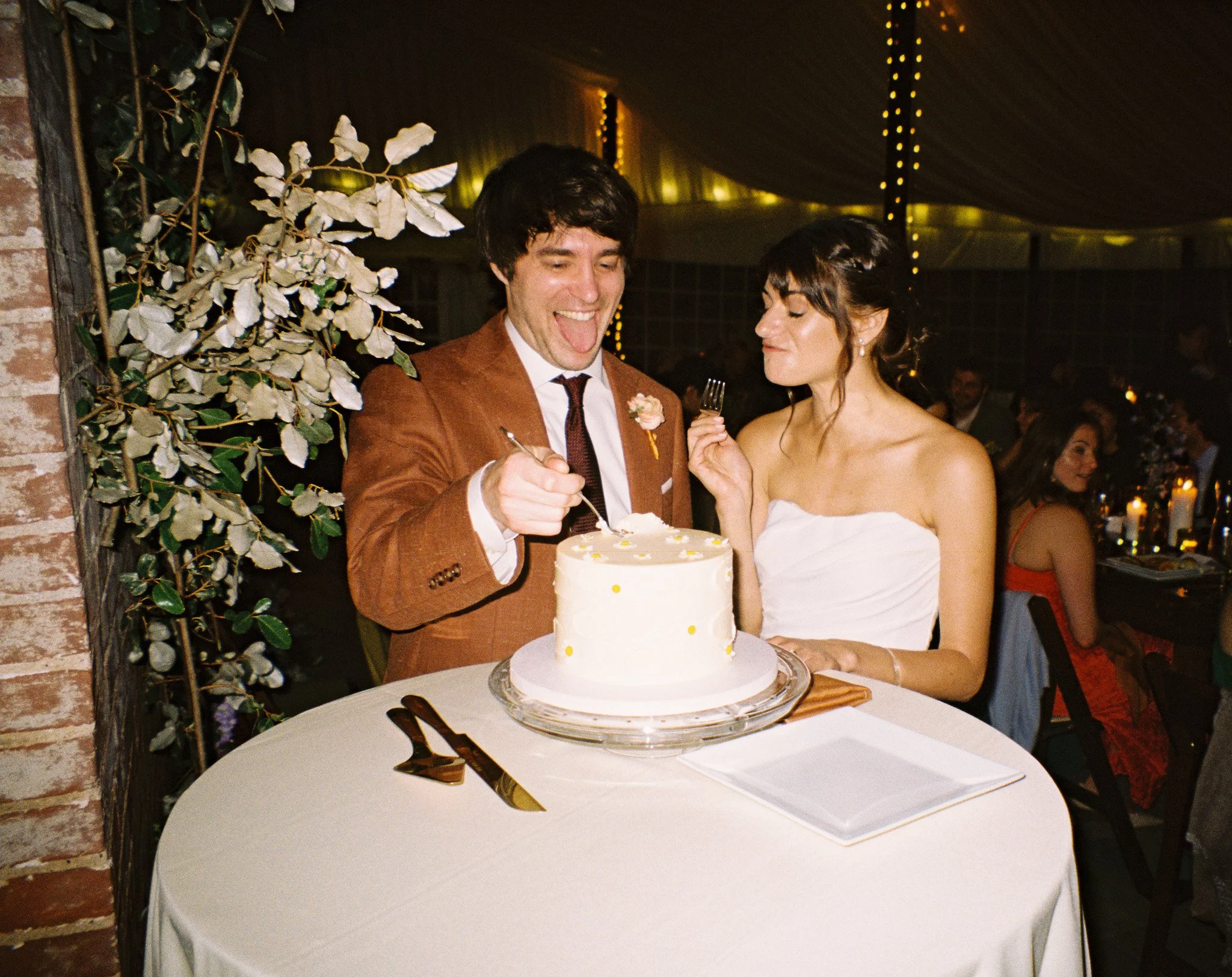 A couple at their wedding reception cutting a white wedding cake with yellow dots, sitting at a round table with a white tablecloth, gold forks, and napkin rings, with wedding guests in the background.