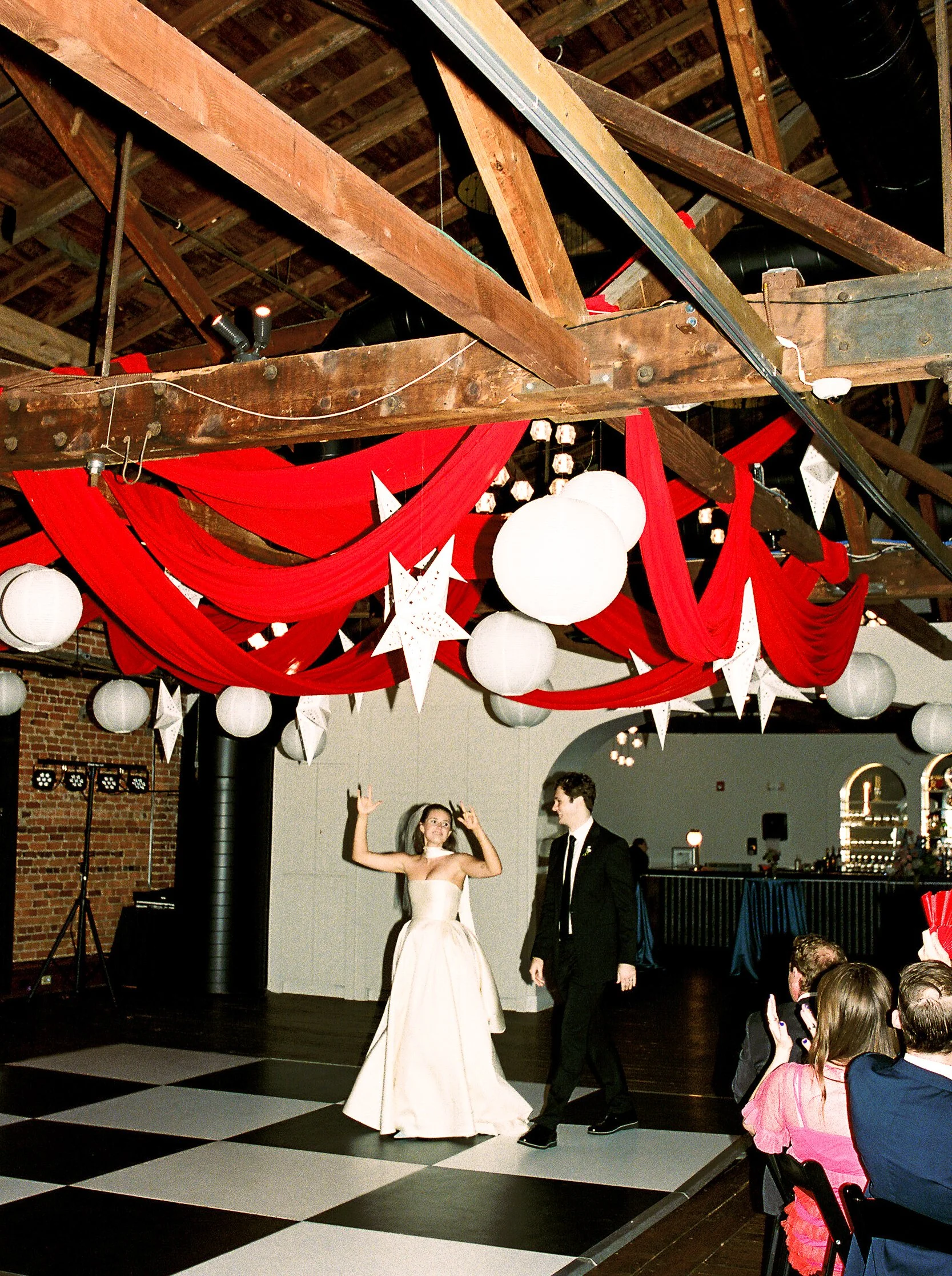 A girl in a white ball gown dancing with a boy in a black suit at a wedding reception in an industrial-style venue decorated with red drapes, white paper lanterns, and hanging white paper stars, with guests watching and taking photos.