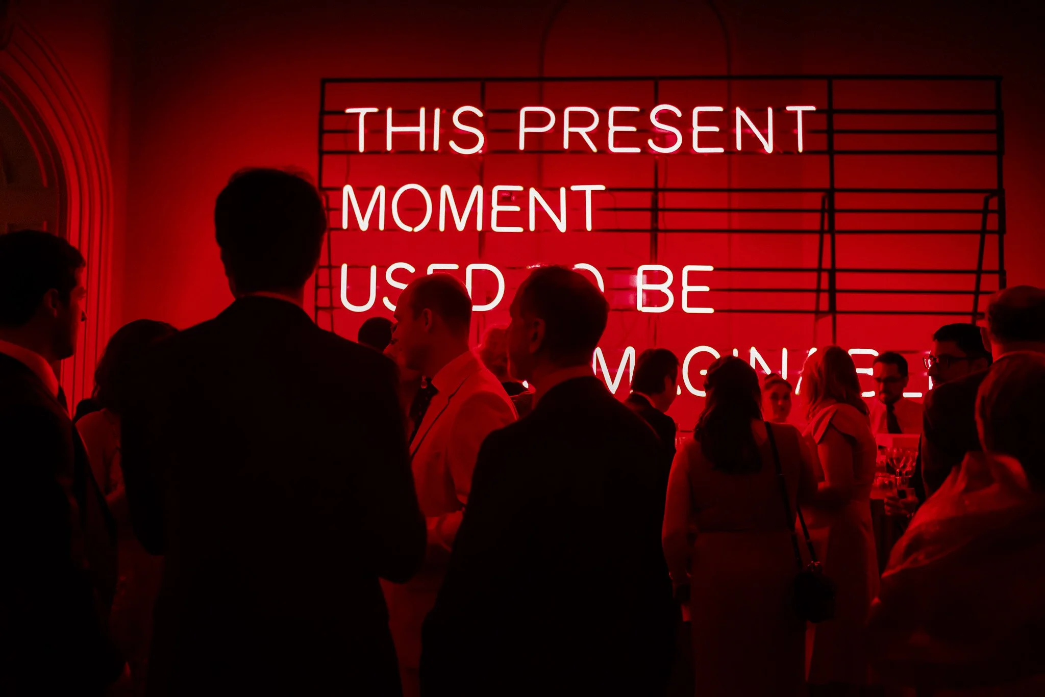 People in formal attire at an event with a bright neon sign that reads 'This Present Moment Used to Be' on a red background.