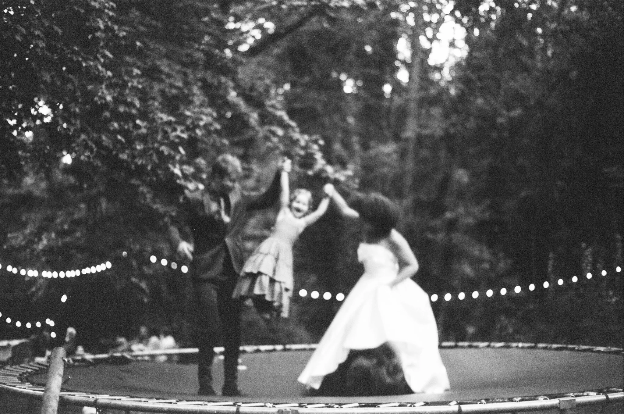 Black and white photo of children playing on a trampoline in an outdoor setting with trees in the background, surrounded by string lights.