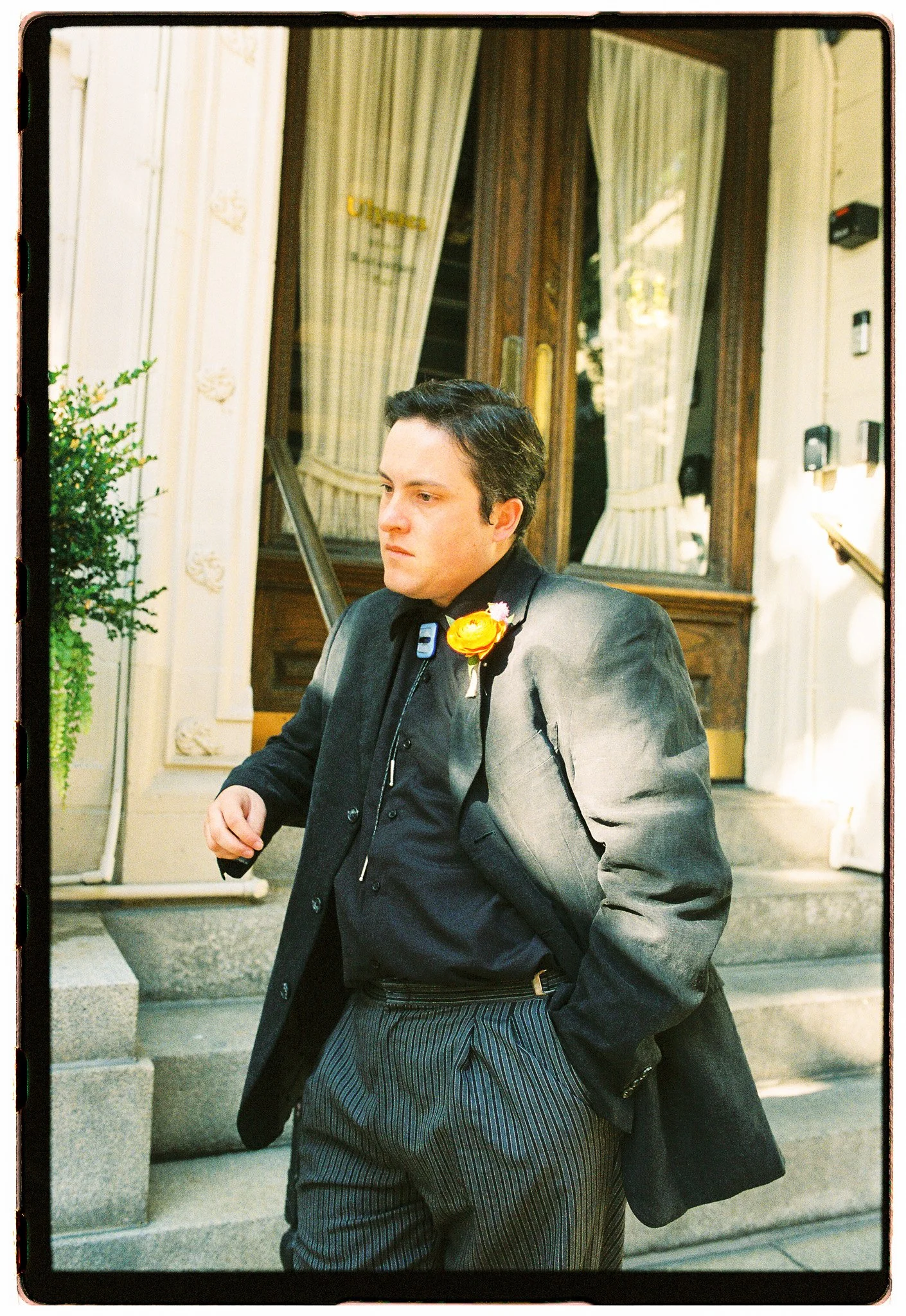 A man in a gray suit with a boutonniere stands outside a building, with a serious expression on his face. The background shows a door with curtains and some greenery.
