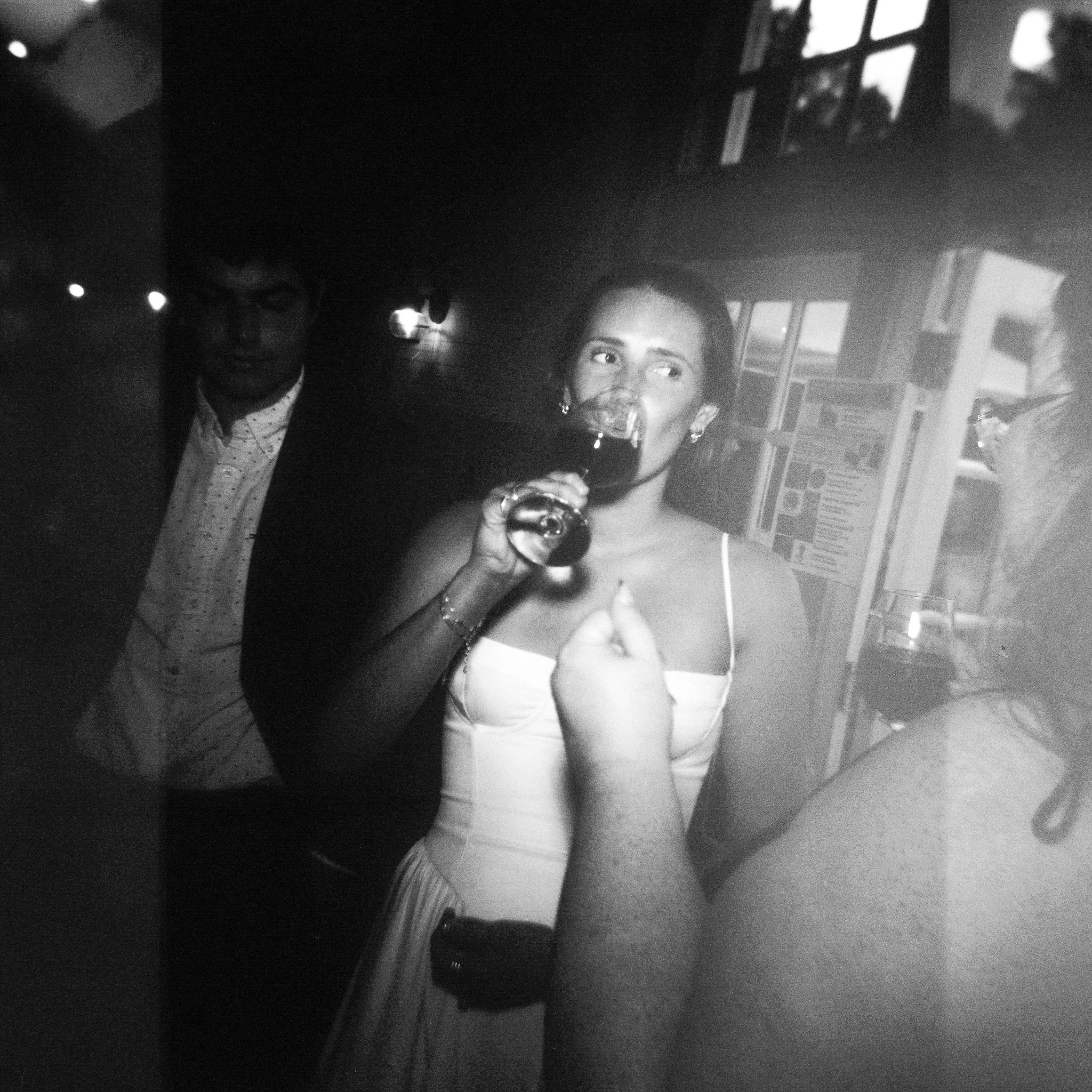 A woman taking a selfie in a mirror while holding a wine glass, with a man in the background and bookshelves visible in the room.