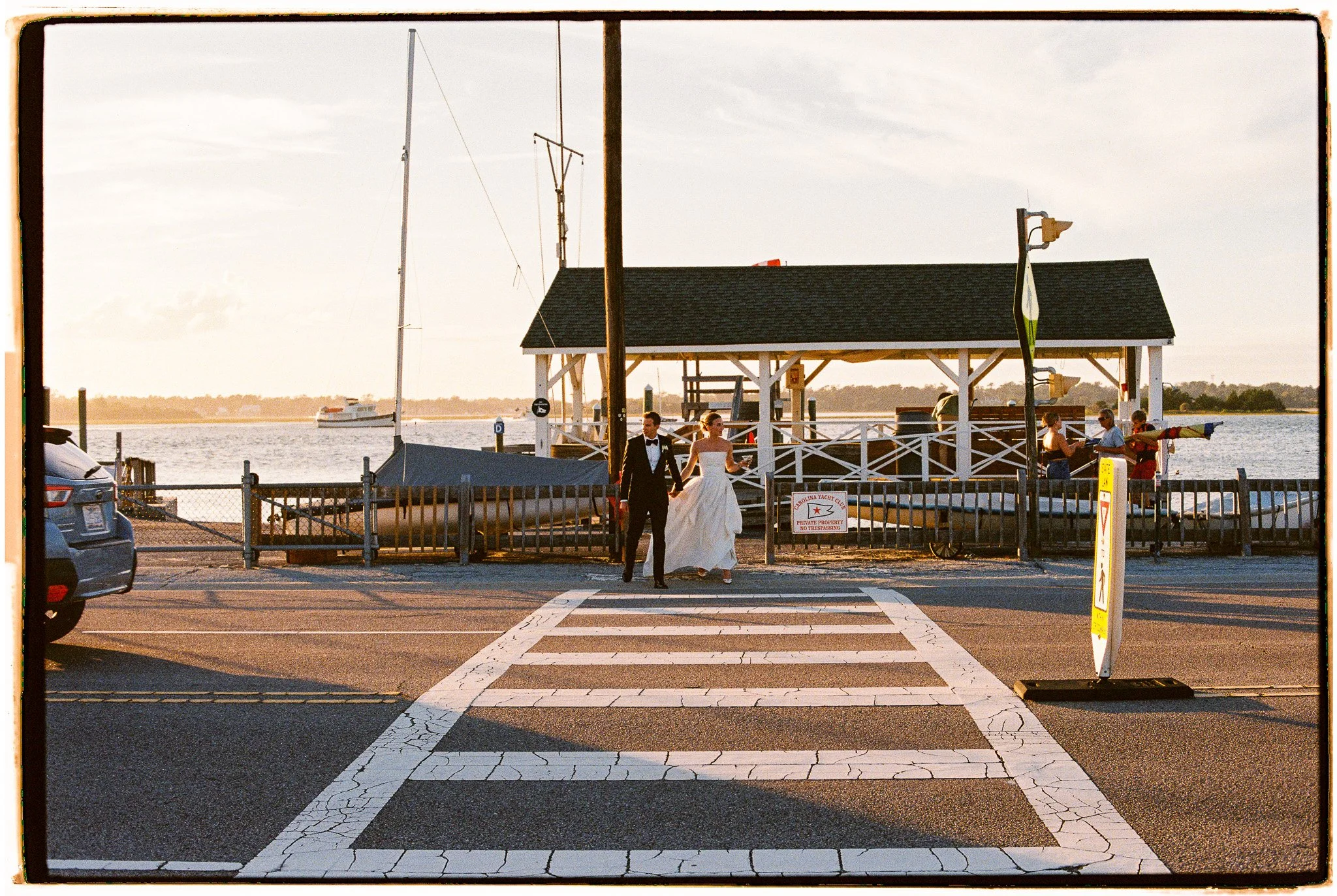 A waterfront scene with a wooden dock, two people in formal attire walking along a pier, a boat on the water, and a sky with scattered clouds.
