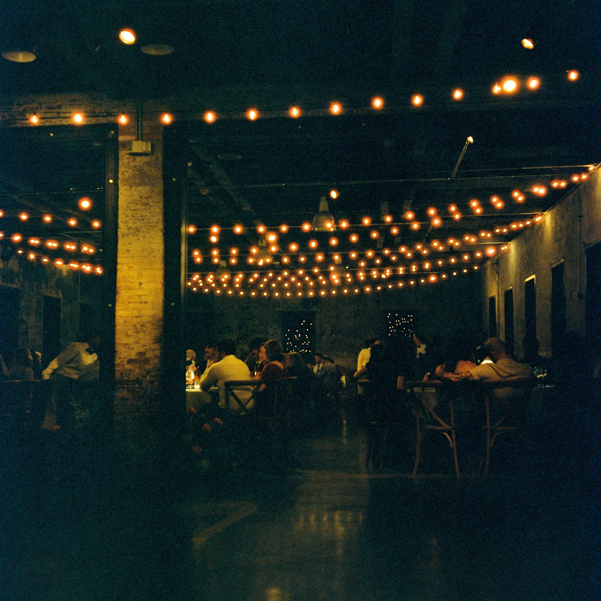 Night scene with hanging string lights illuminating an outdoor patio area filled with tables and people.