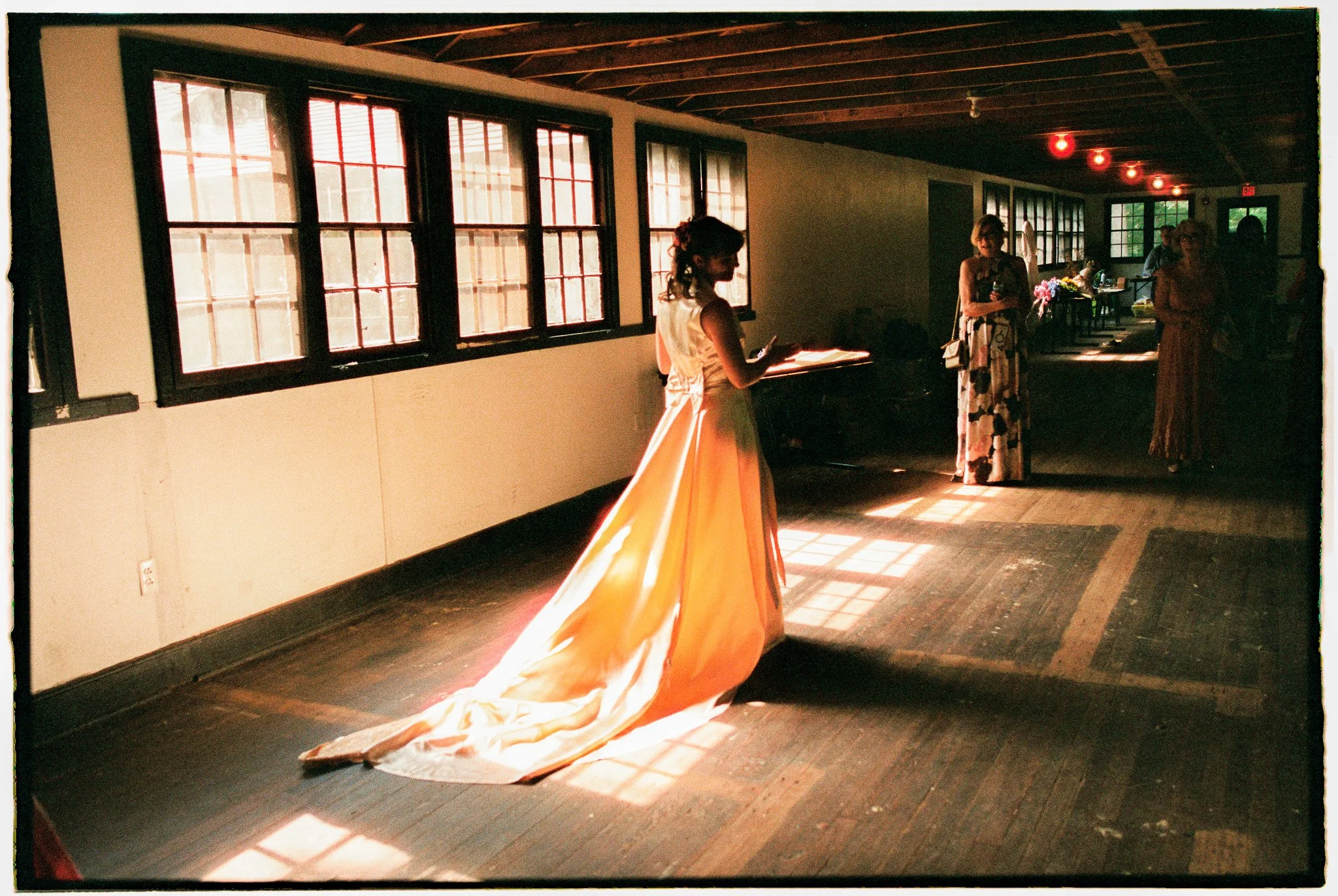 A woman in a white gown with a long, flowing orange train standing in a room with large windows and wooden floors, while other women in dresses are near her; the lighting suggests it is daytime.