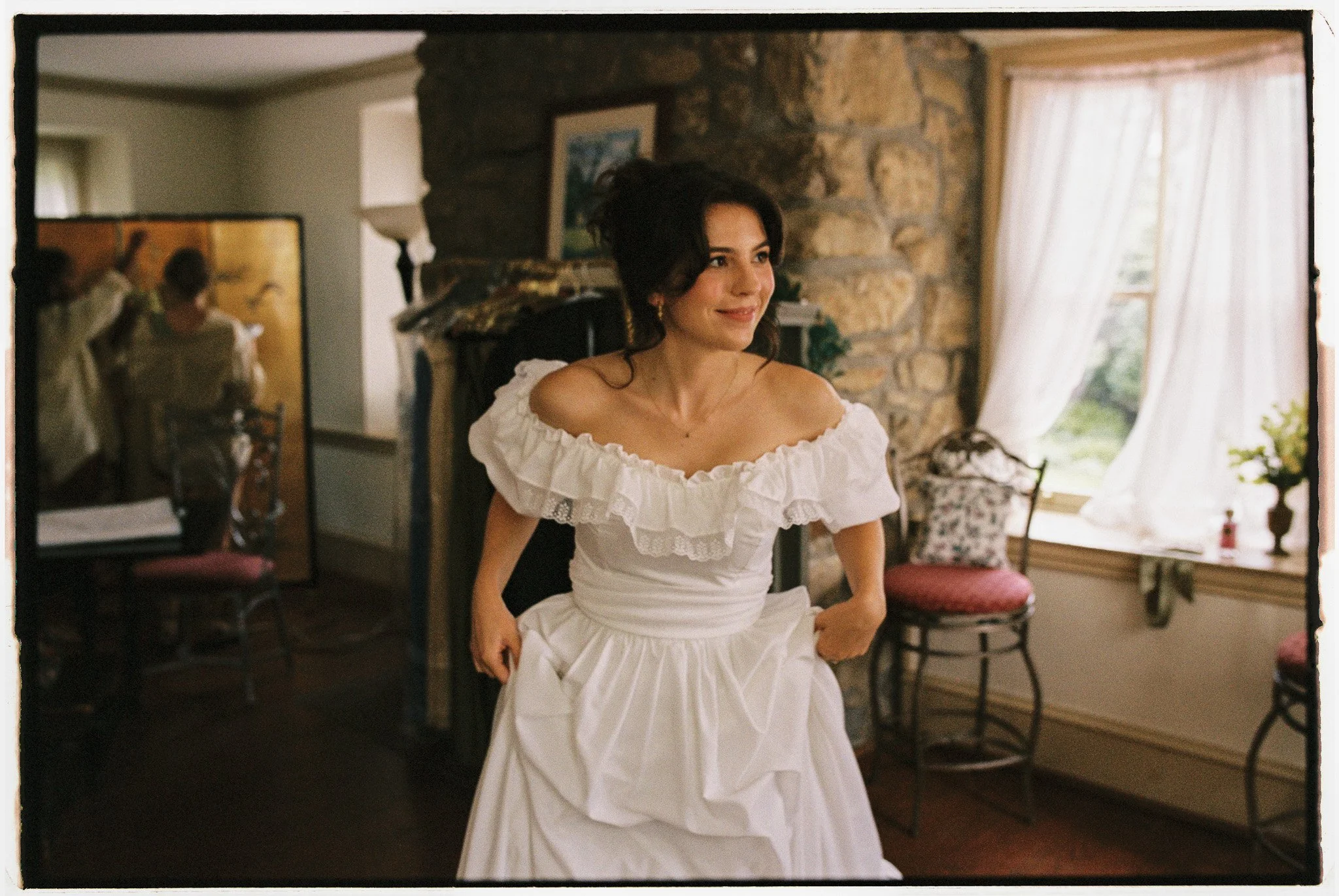 Woman wearing a white dress, smiling, inside a cozy room with stone walls and large windows with white curtains.
