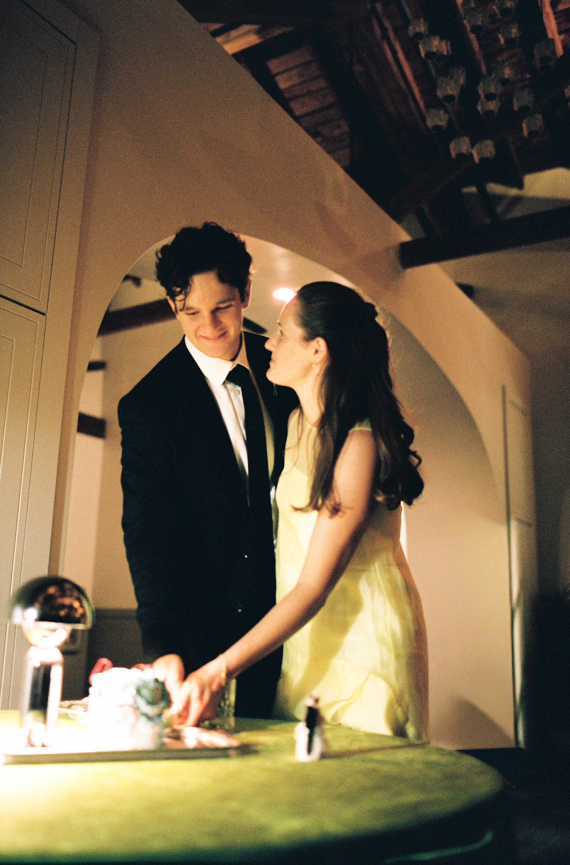 A young man in a black tuxedo and a woman in a yellow dress sharing a moment during a celebration, possibly a birthday or special event, with a cake in front of them.