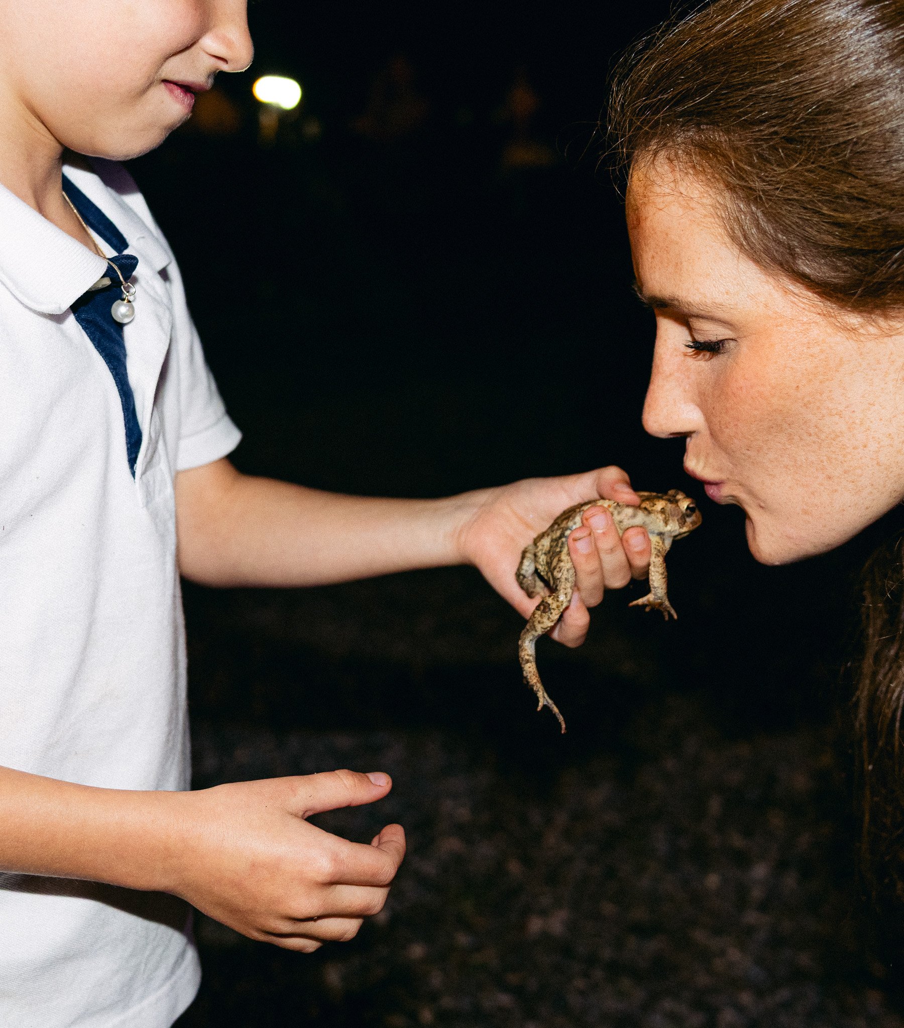 A young girl holding a lizard with another girl in a white shirt standing close by at night.