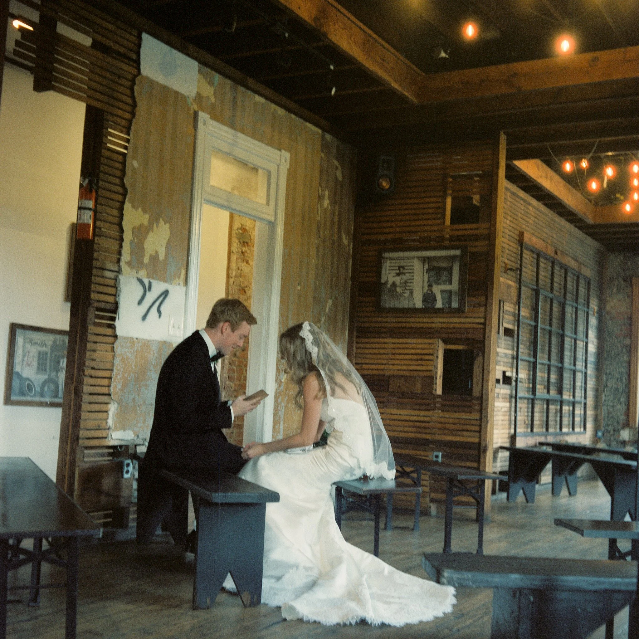 A couple getting married at an indoor wedding ceremony, with the bride in a white gown and veil and the groom in a black suit, standing before a rustic wooden backdrop. The backdrop features a map and artwork, illuminated by string lights. A person t
