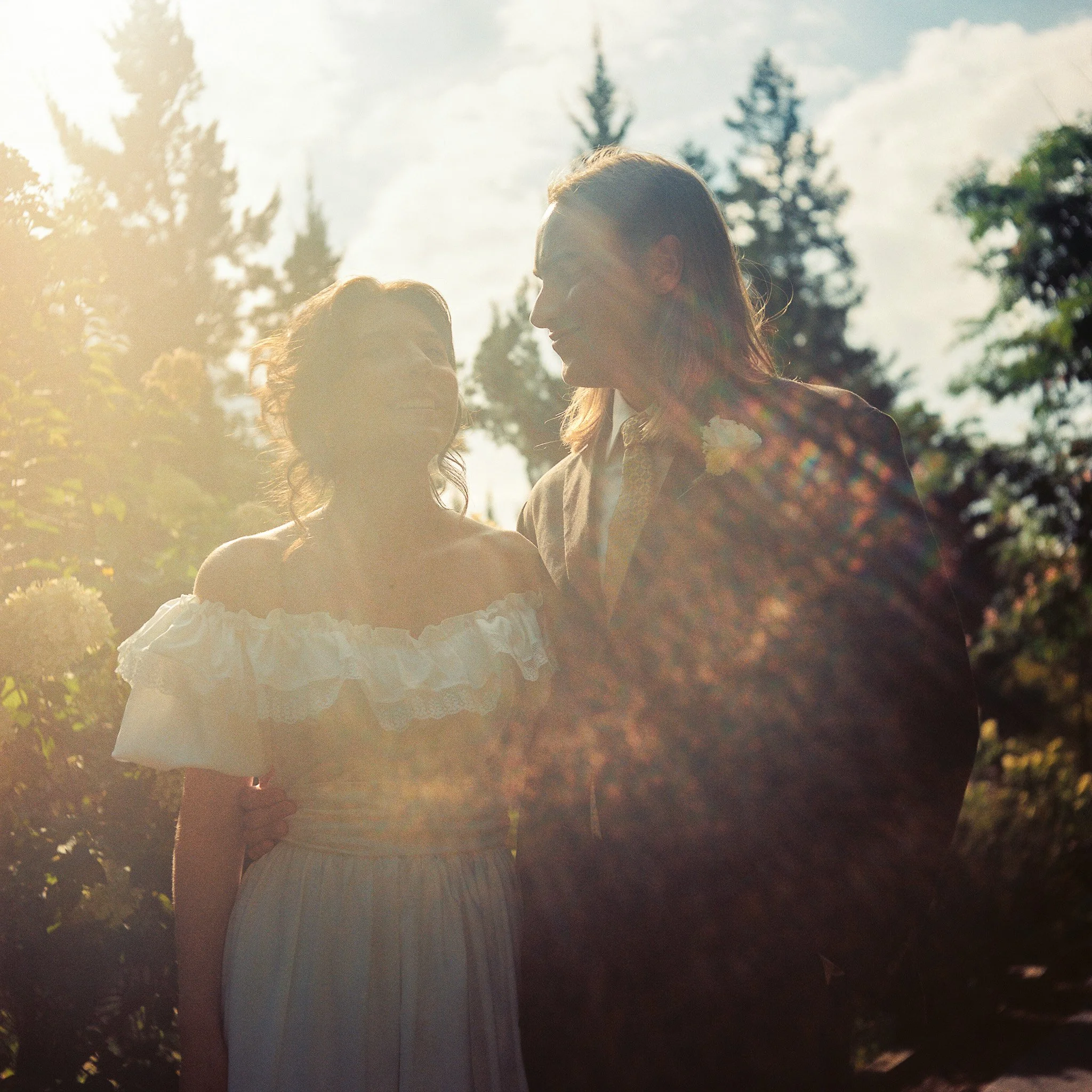 Two women, one with red hair and one with dark hair, in an outdoor setting with trees and a partly cloudy sky, sharing an intimate moment.