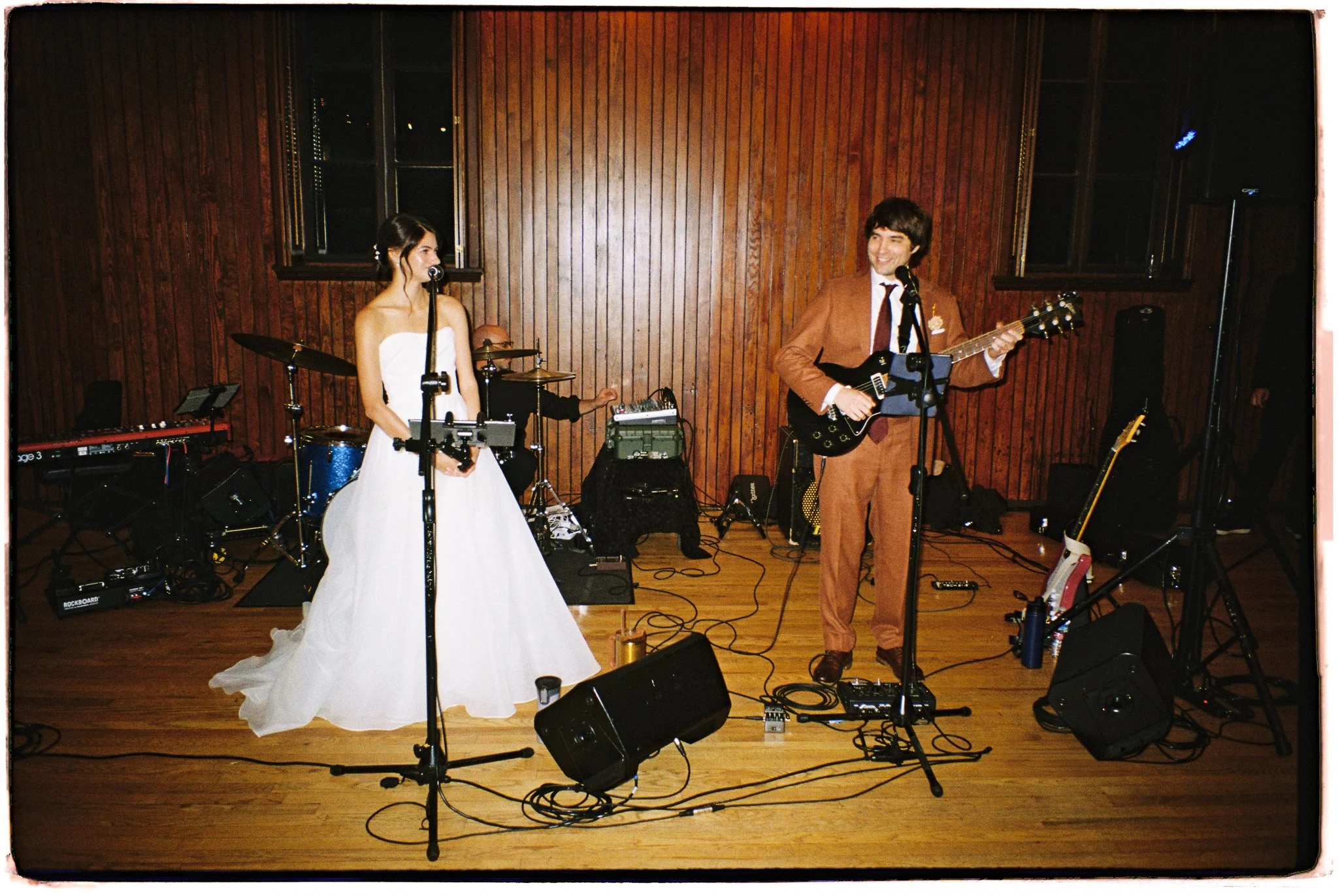 A band performing on stage with a female singer in a white dress and a male guitarist in a brown suit, with musicians and equipment in the background.