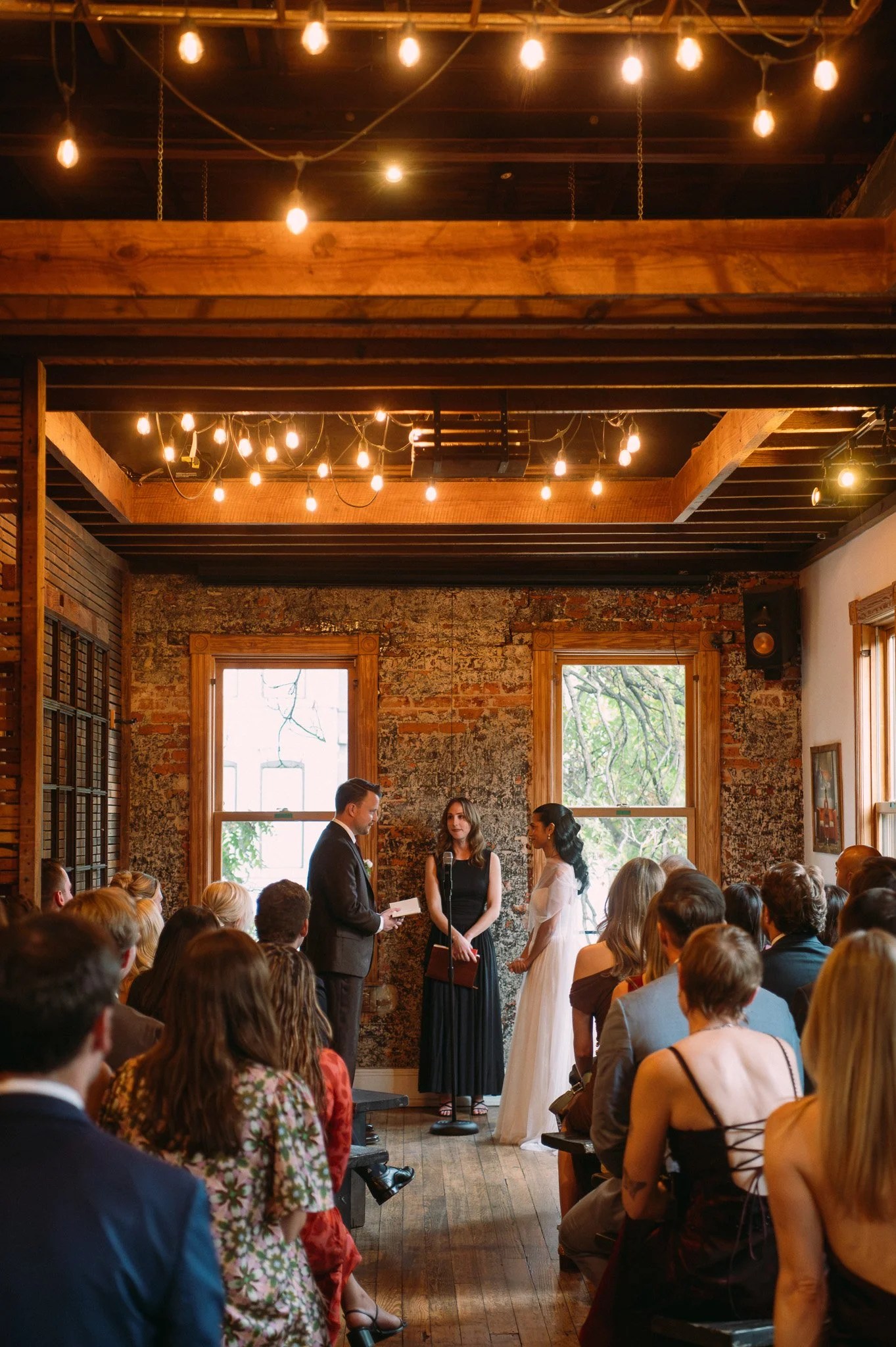 A wedding ceremony taking place indoors with a rustic, exposed brick wall, large windows, and hanging string lights. A couple is standing before an officiant, exchanging vows, with guests seated and observing.