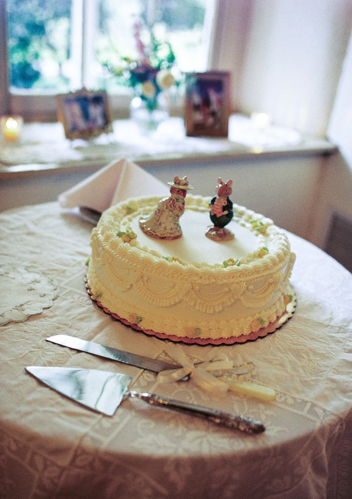 A decorated cake with two animal figurines on top, placed on a table with a lace tablecloth. The background shows a window with flowers and framed pictures.