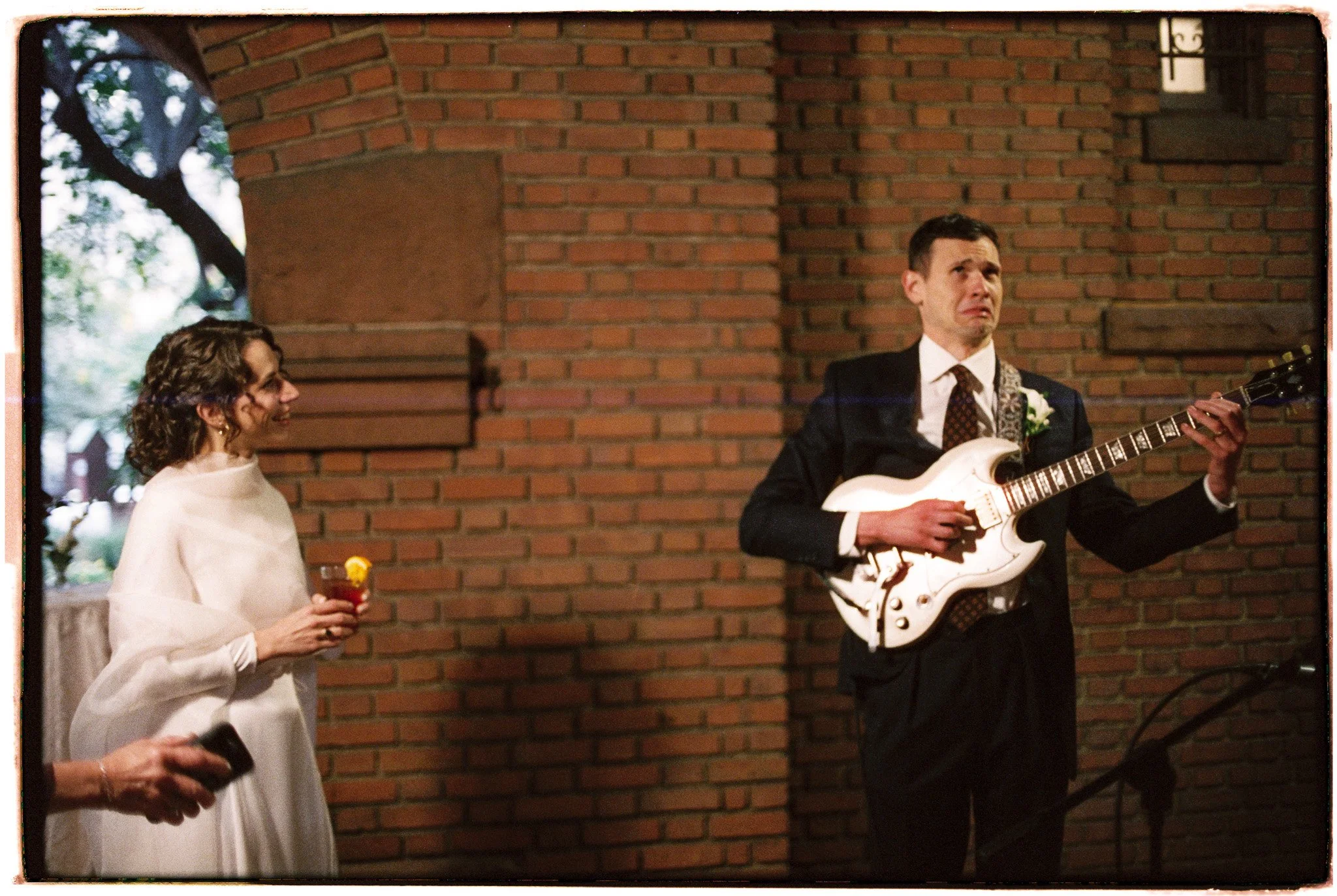 A man in a suit playing an electric guitar at a gathering, and a woman in a white dress holding a drink, standing against a brick wall.