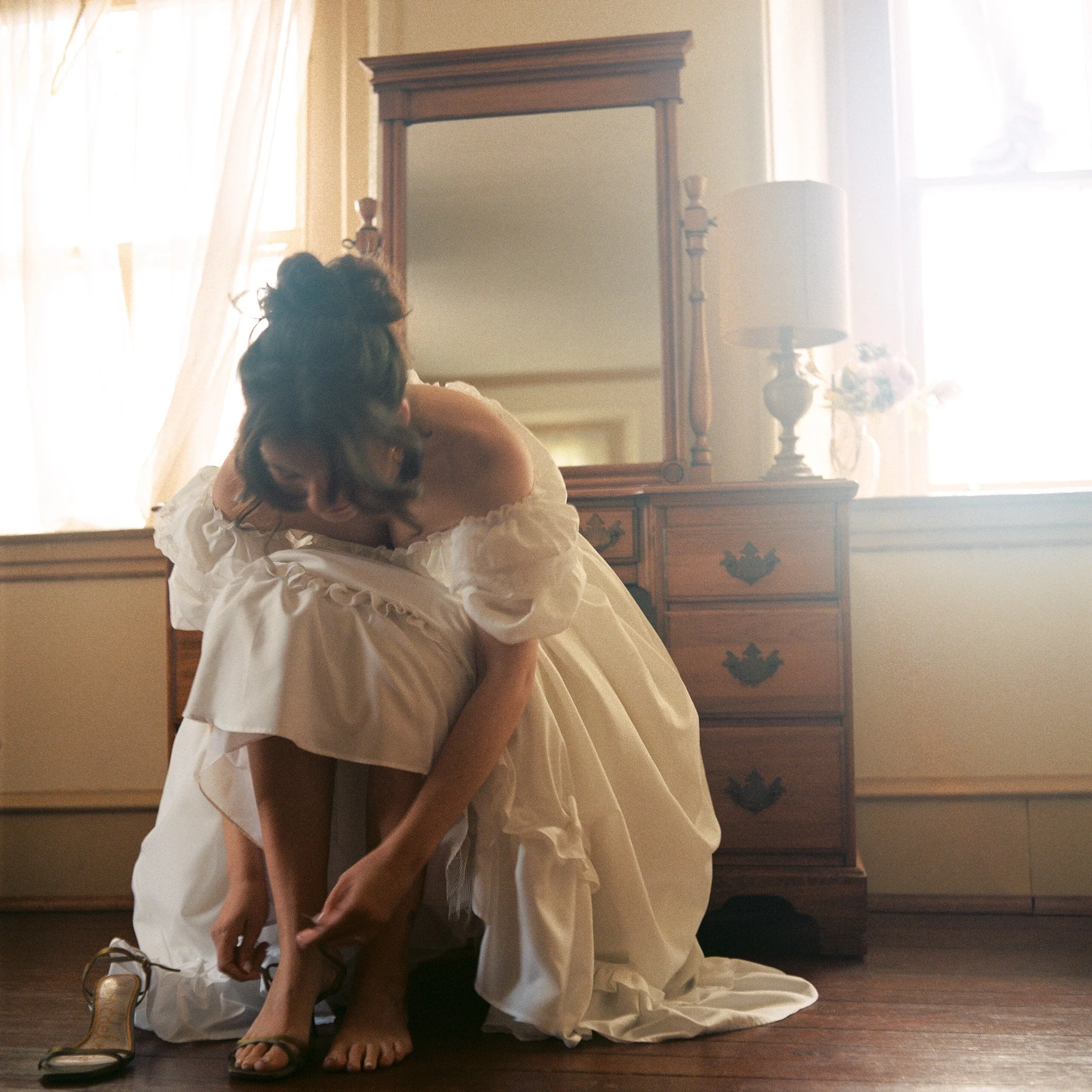 A woman in a white dress is sitting on the floor, adjusting her gold high-heeled sandal. She has dark, curly hair and is near a wooden dresser with a large mirror and a lamp. The room is softly lit with natural light coming through sheer curtains.