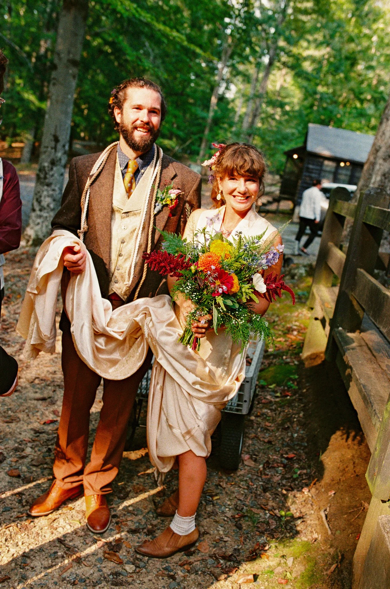 A smiling man and woman walking outdoors on a sunny day. The woman holds a colorful bouquet of flowers. The man is dressed in a vintage-style brown jacket and pants, with a shirt and tie, and the woman wears a cream-colored dress with puffy sleeves a