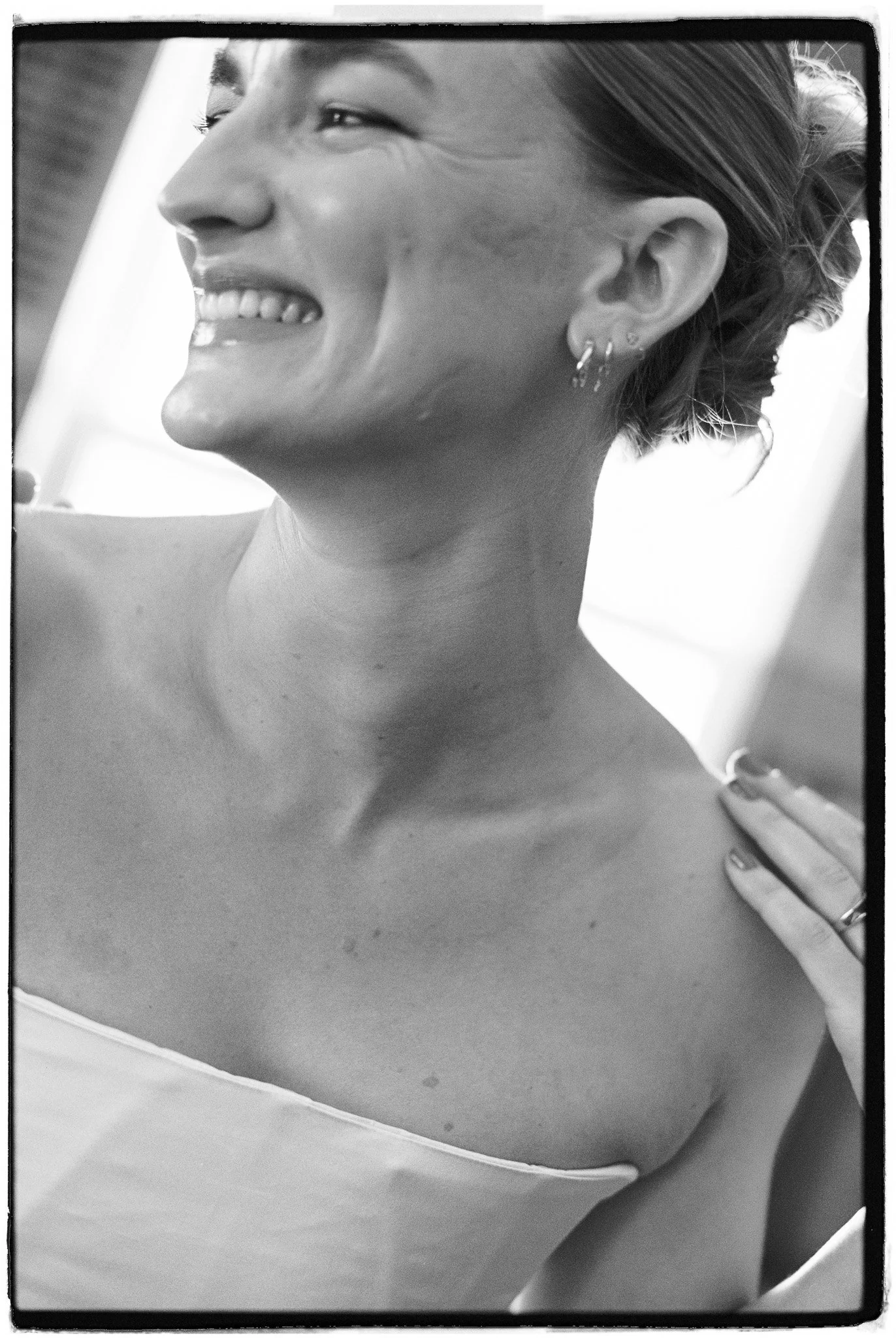Close-up black and white photo of a woman smiling, showing her teeth, with earrings, her short hair styled back, her face turned slightly to the side, and her hand resting on her shoulder.