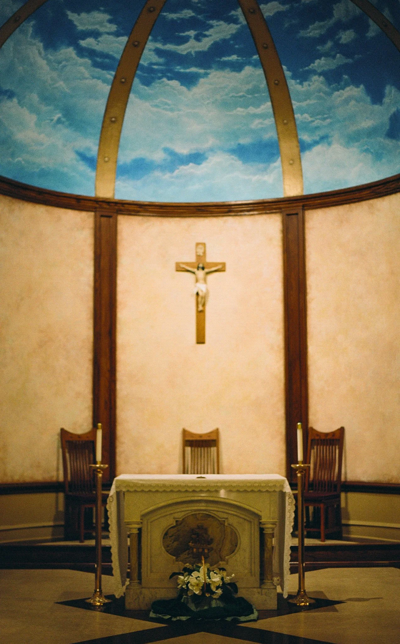 Interior of a church altar with a crucifix, four chairs, candles, and a floral arrangement, with a sky painted on the ceiling.