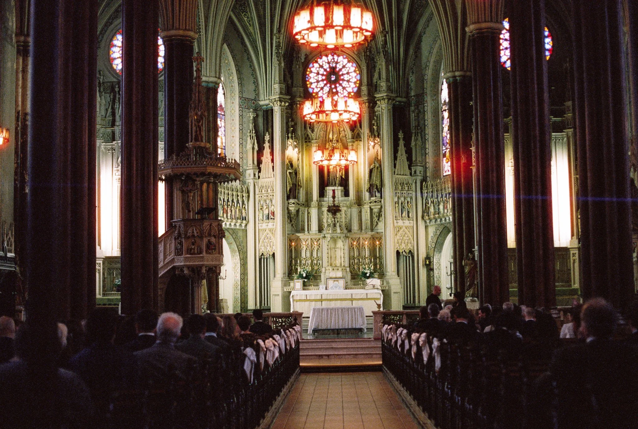 Interior of a church with high vaulted ceilings, stained glass windows, large red chandeliers hanging from the ceiling, and rows of pews filled with people attending a service or event.