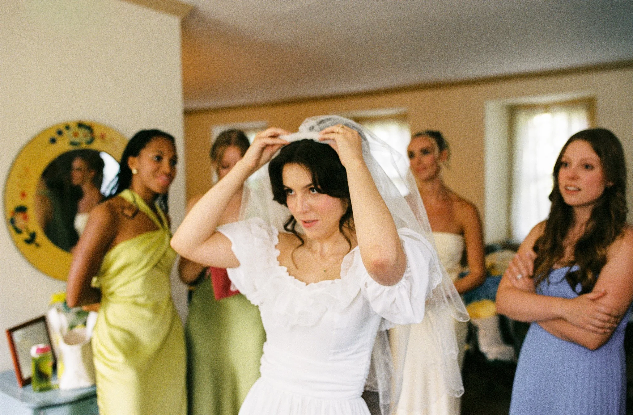 A woman in a white dress is putting on a veil with the assistance of other women in a room with beige walls and a window.