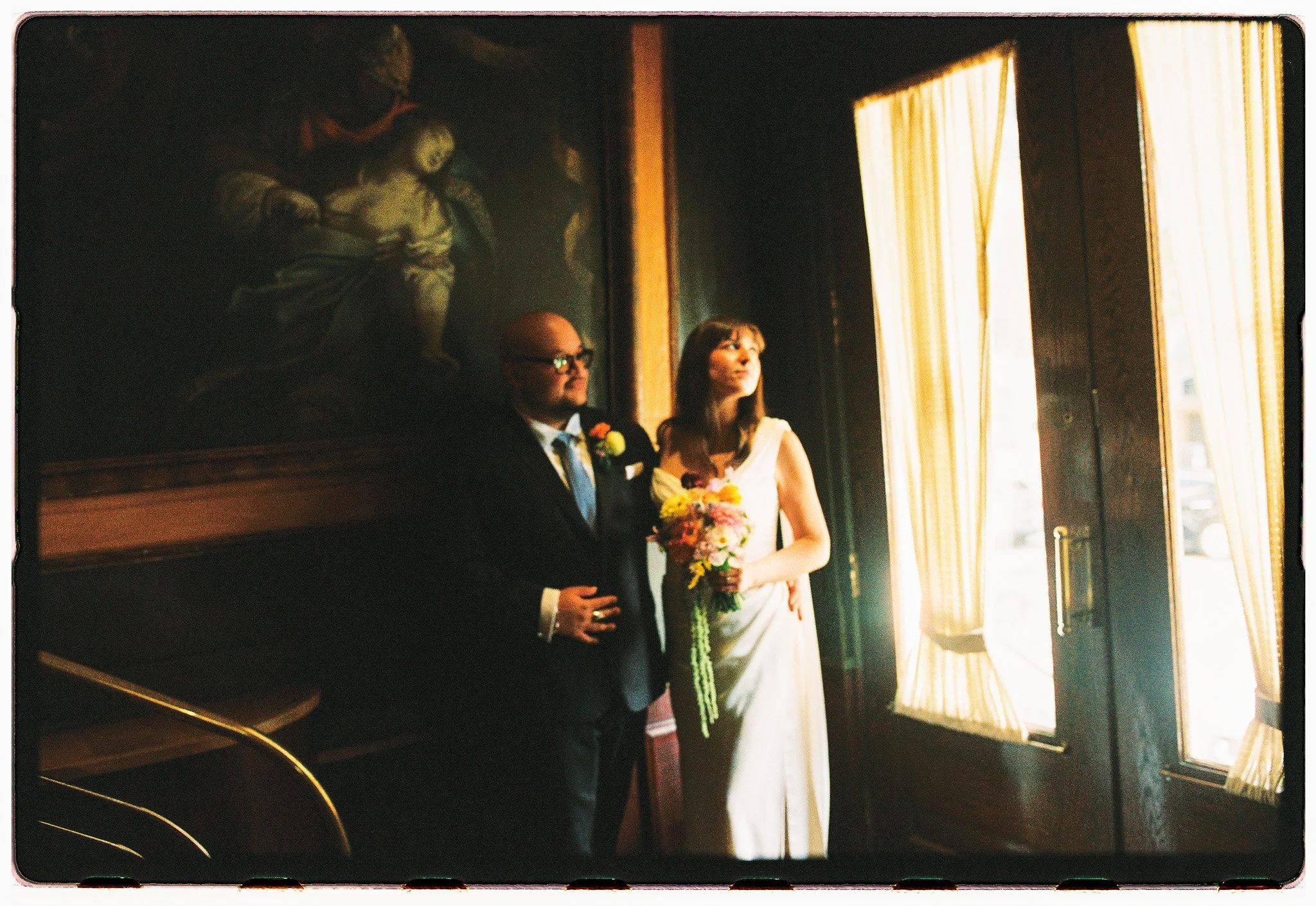 A man in a suit and a woman in a white dress with a floral bouquet inside a room with gold curtains and large paintings on the wall.