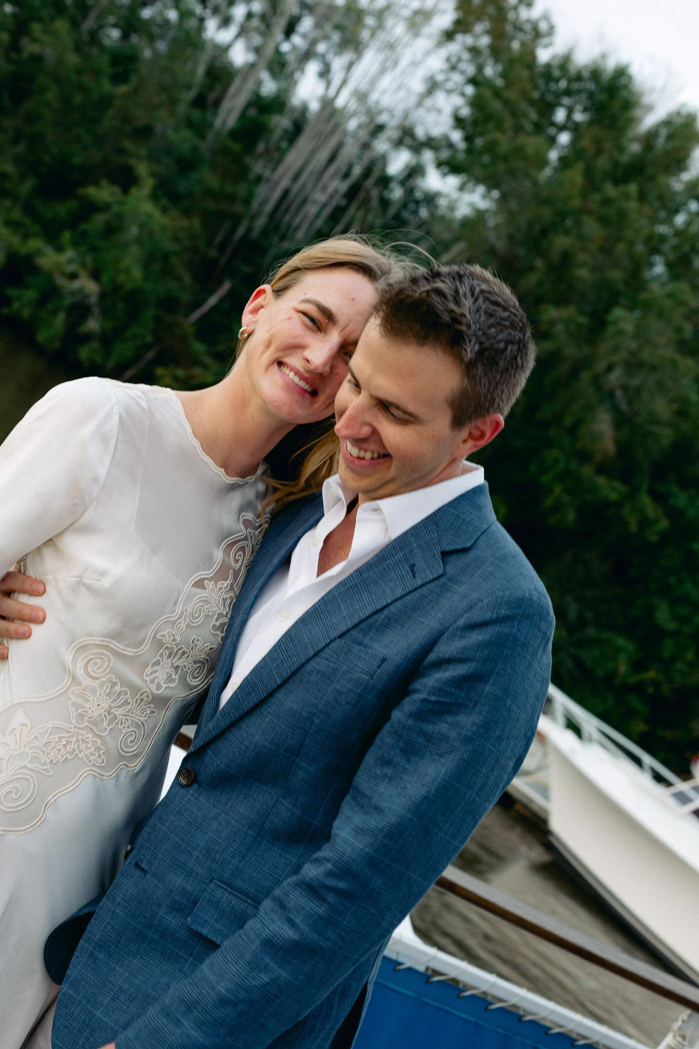 A smiling woman in a white dress with floral embroidery and a man in a suit with a checkered pattern, both leaning their heads together outdoors in front of a green, tree-filled background.