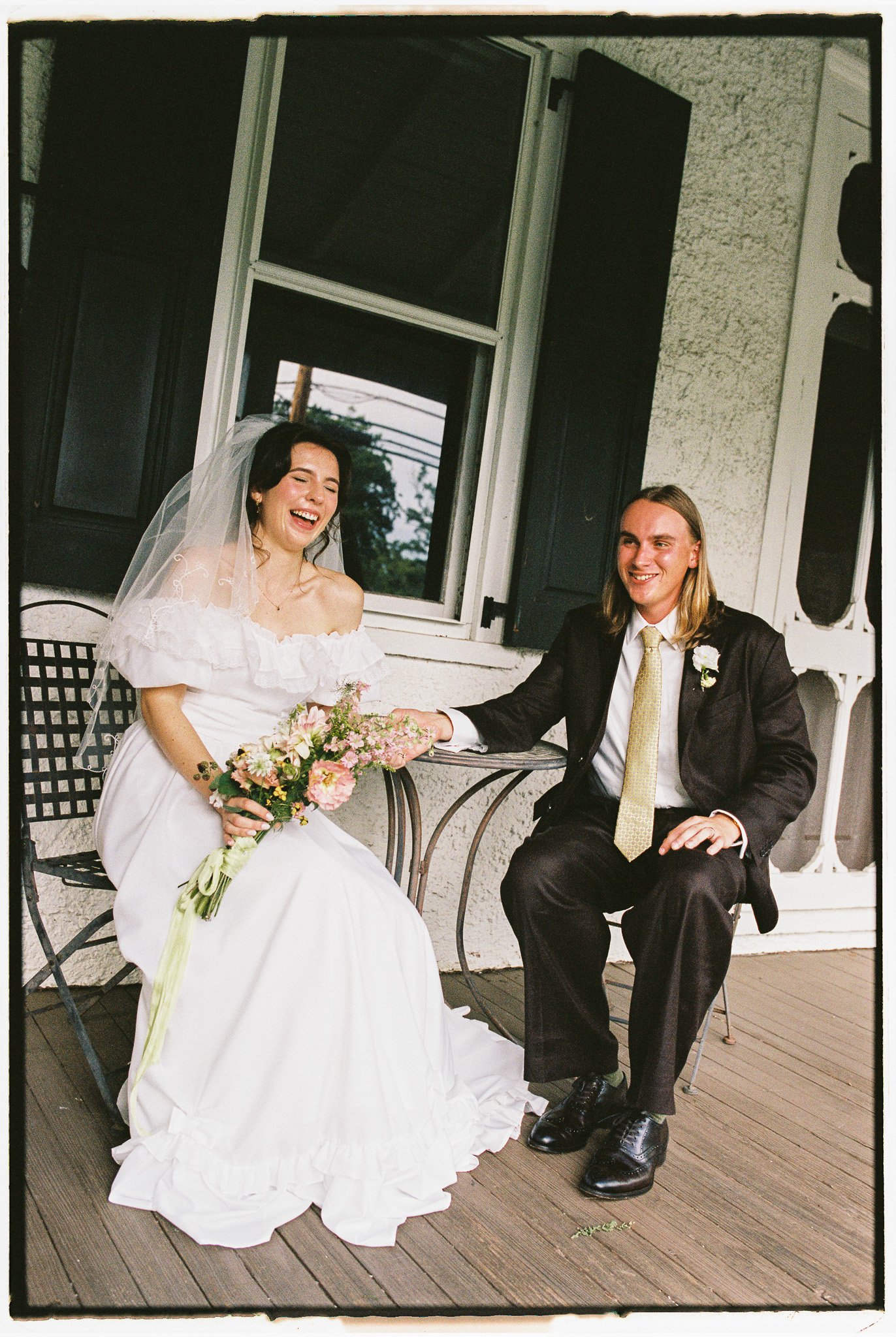 A bride and groom are sitting outside, holding hands and smiling. The bride is wearing a white wedding dress with an off-shoulder neckline and a veil, and holding a bouquet of flowers. The groom is dressed in a black suit with a white shirt, yellow t