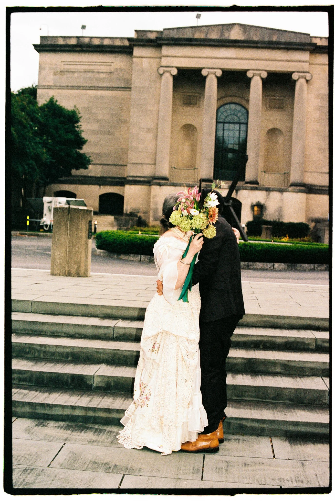 A couple embracing on the stairs outside a building, with the woman holding a bouquet of flowers in front of her face, obscuring her face. The building has large windows and tall columns.