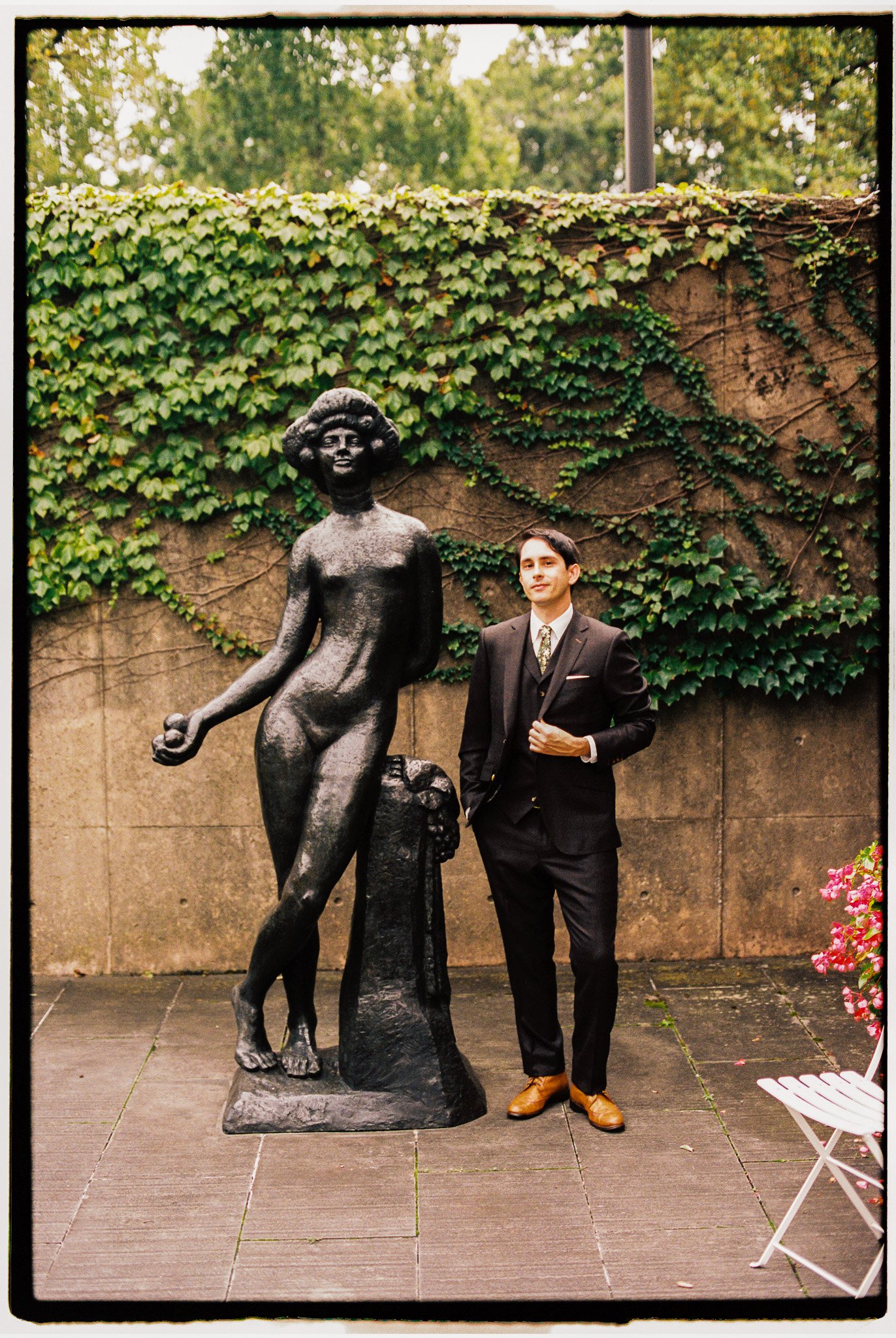 A man in a black suit and brown shoes standing next to a large bronze statue of a woman with short hair. The background features a stone wall covered with green ivy and other plants, with trees and an overcast sky visible above.