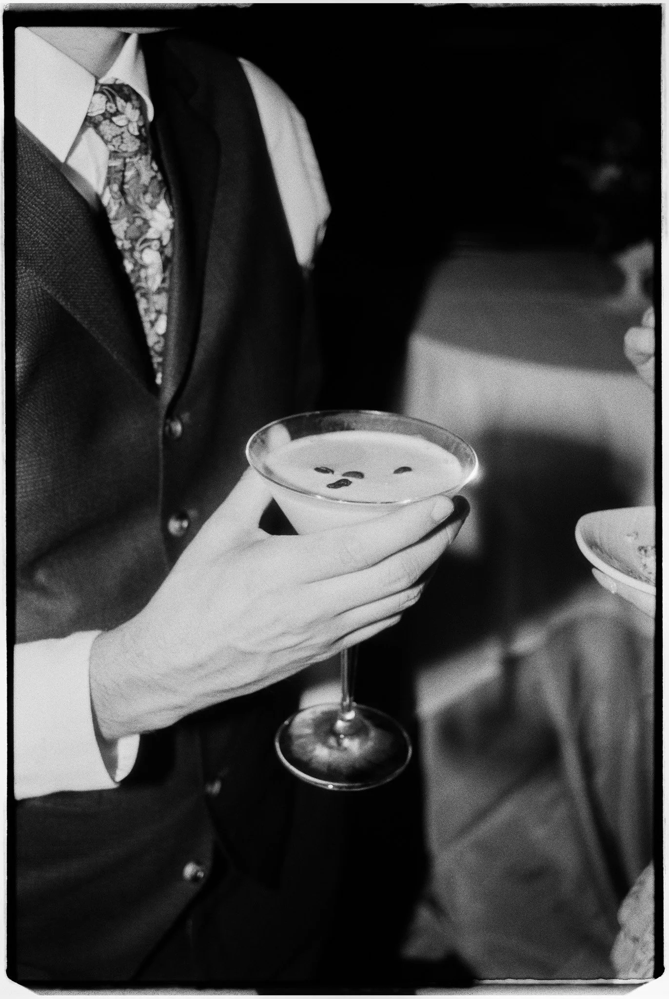 A man dressed in a suit with a floral patterned tie holds a martini glass with an olive garnish.