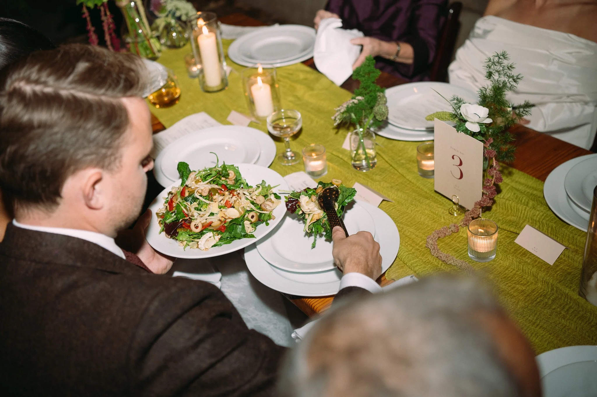 A man in a brown jacket is holding a plate of salad at a decorated dinner table with candles and floral arrangements.