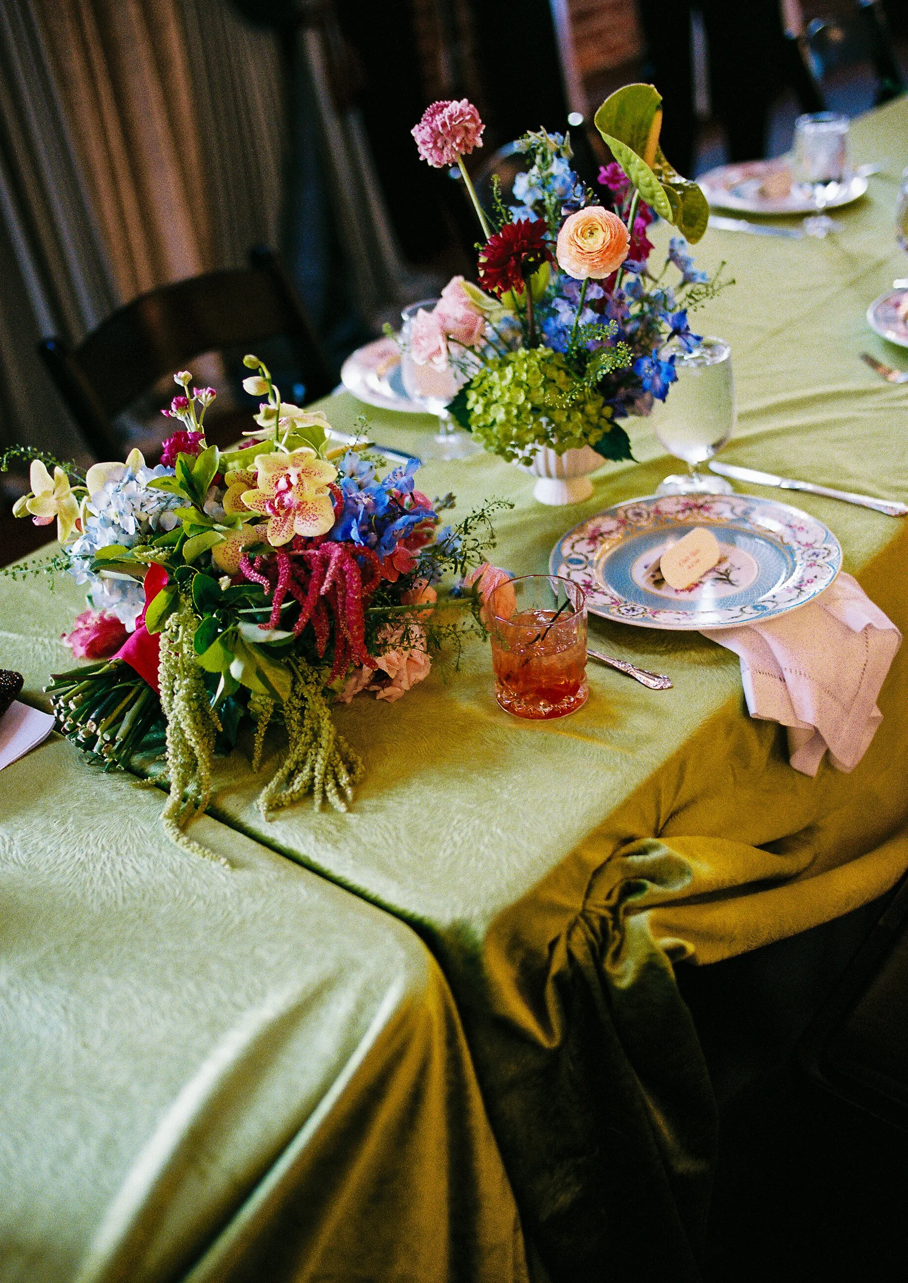 A table set with floral centerpieces, plates, and glasses on a green tablecloth and yellow tablecloth. The flowers are in various colors including pink, red, blue, and green.