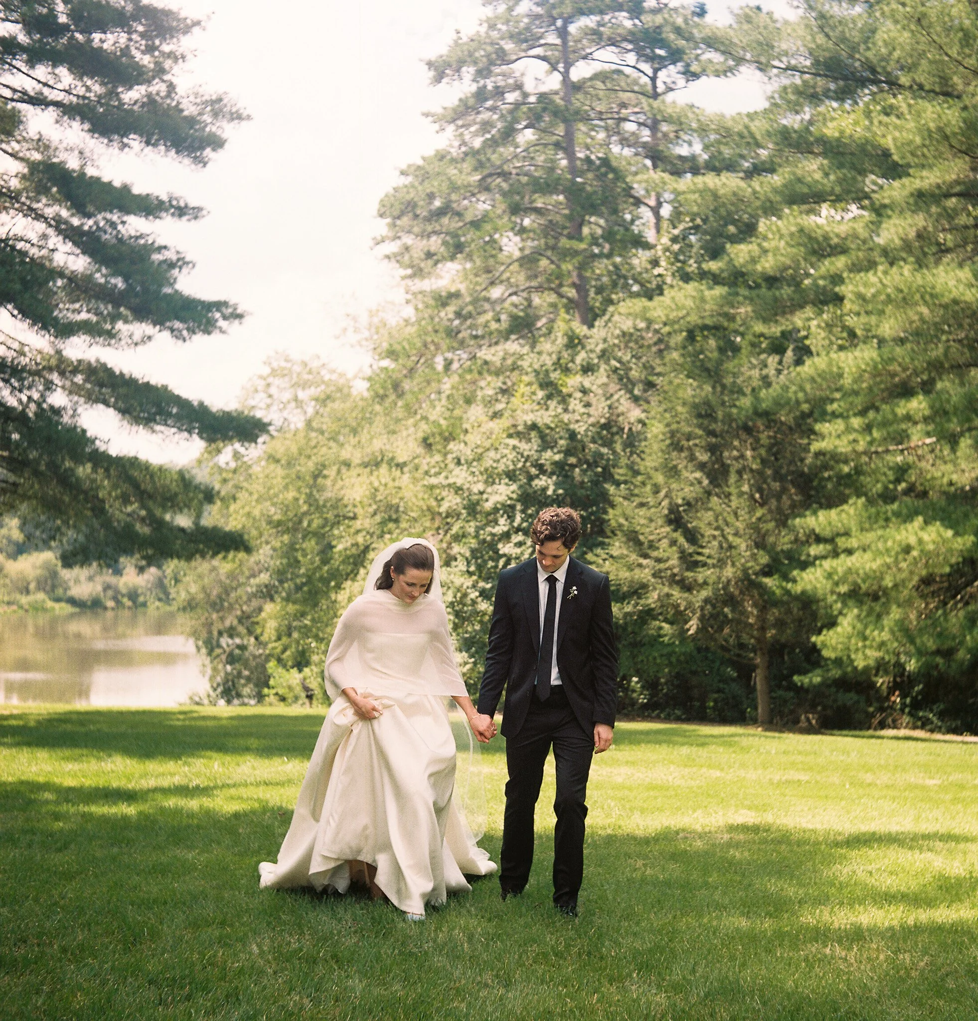 A bride and groom walking hand in hand through a park with green grass and tall trees.