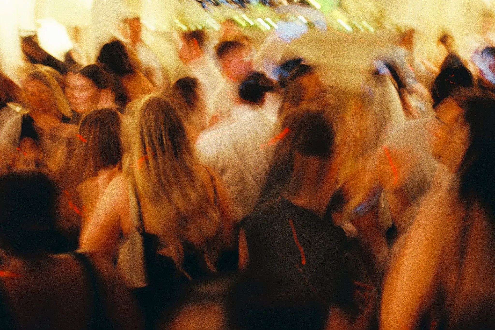 Blurred photo of a group of people at a social event, possibly a wedding or party, with warm lighting and blurry details.