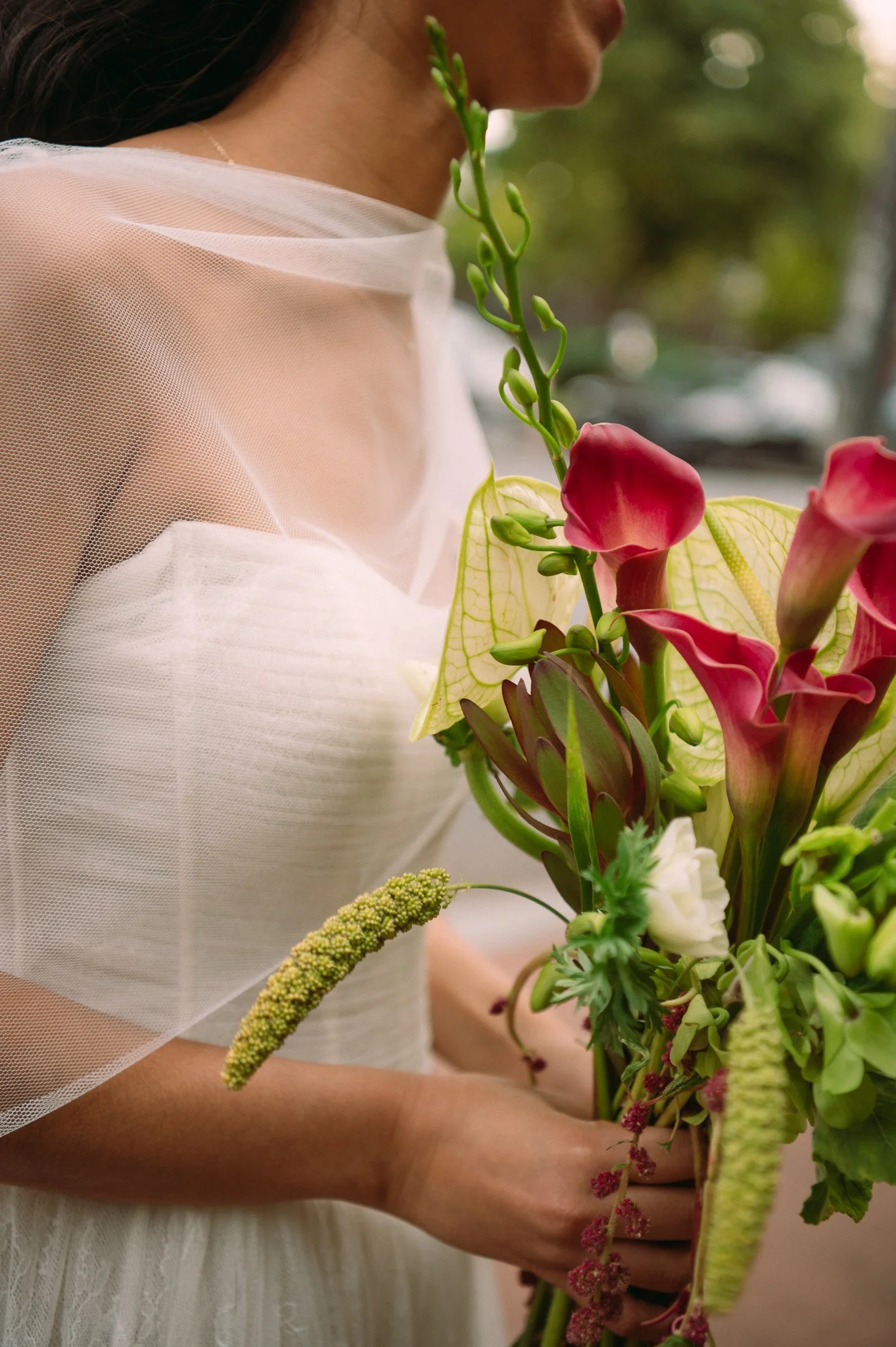 A woman in a white dress holding a bouquet of colorful flowers, including pink and white blooms and greenery, outdoors with blurred trees in the background.