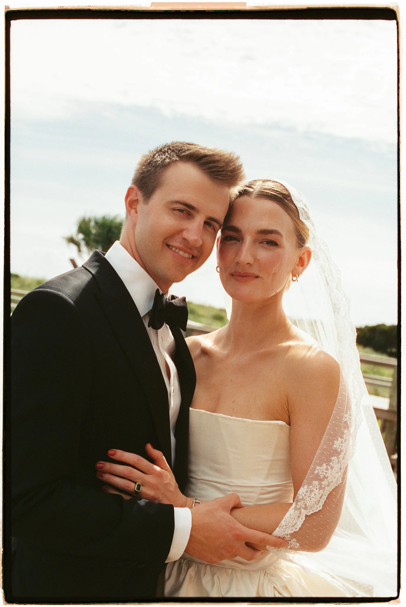 A bride and groom dressed in wedding attire, standing outdoors, smiling and looking at the camera, with a cloudy sky and greenery in the background.